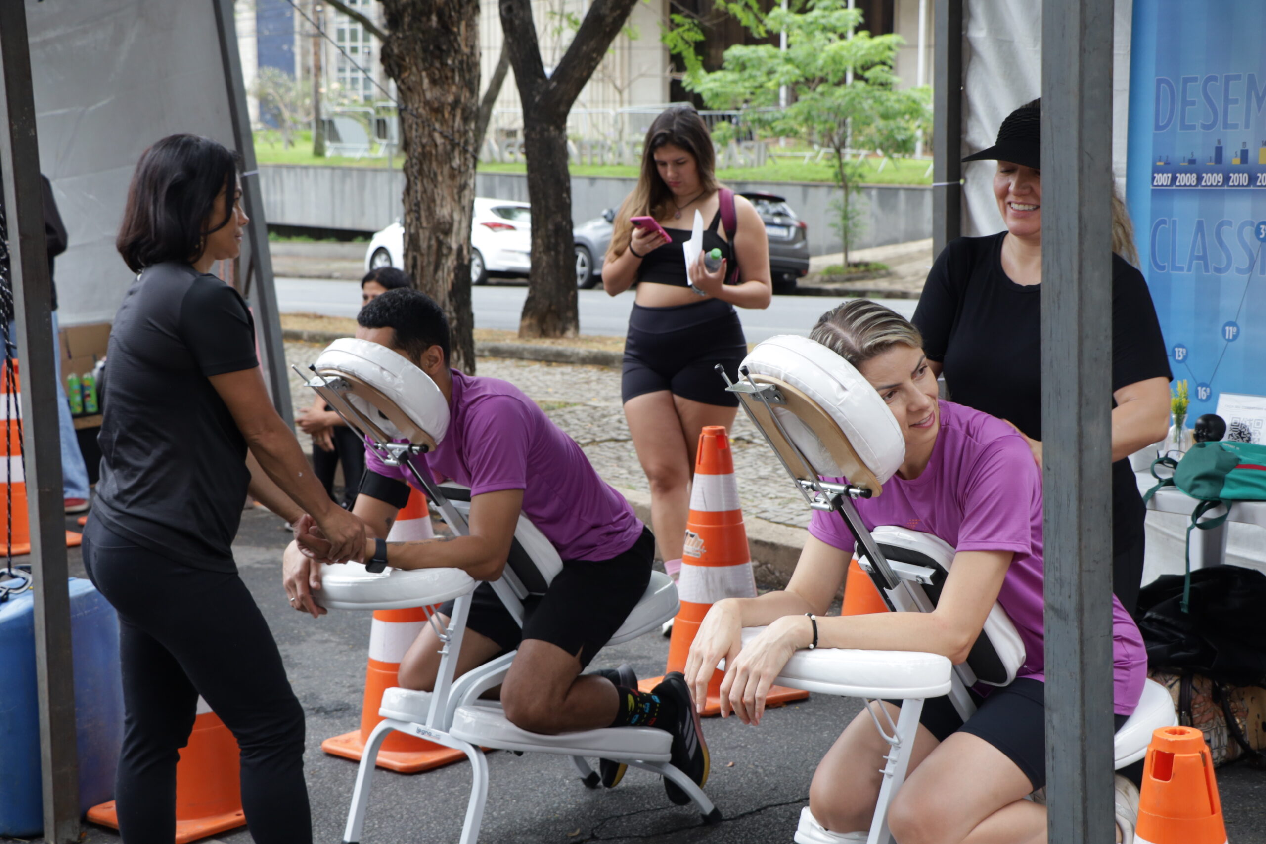 Duas pessoas em camisetas roxas recebem massagem pós-corrida em cadeiras portáteis, em uma área de tenda, com outras pessoas ao redor.