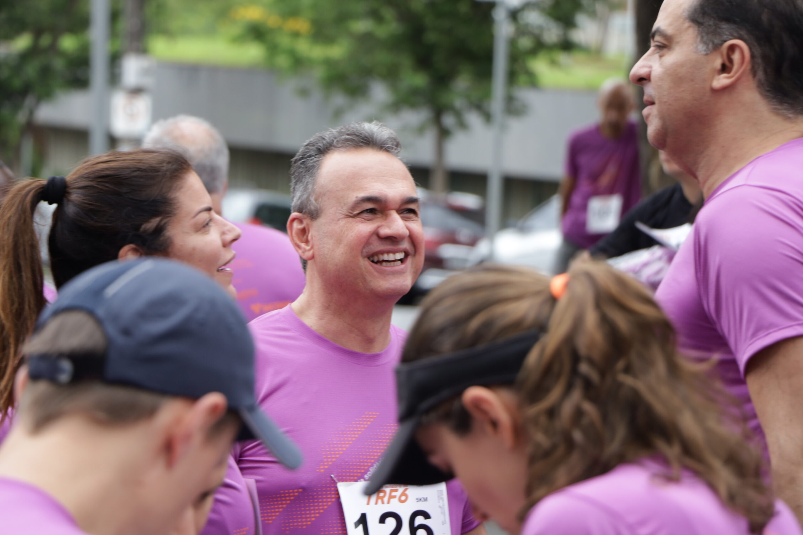 Homem de meia-idade, sorrindo e vestindo camiseta roxa de corrida com número de peito, conversa em grupo após o evento.