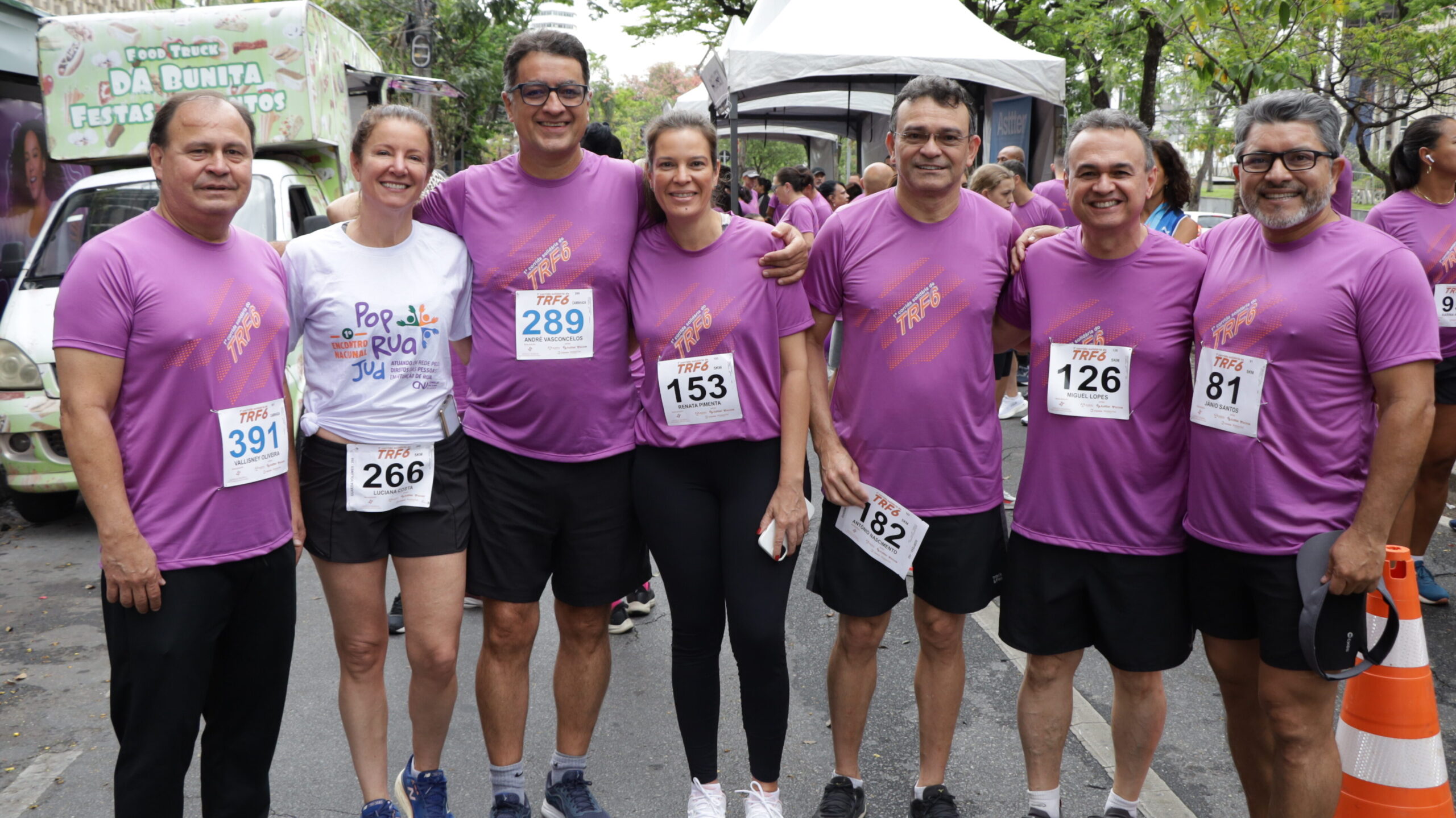 Sete pessoas (cinco homens e duas mulheres) sorriem, vestindo camisetas roxas de corrida (exceto uma mulher de camiseta branca), com números de peito, em um evento de rua com tendas e um food truck ao fundo.