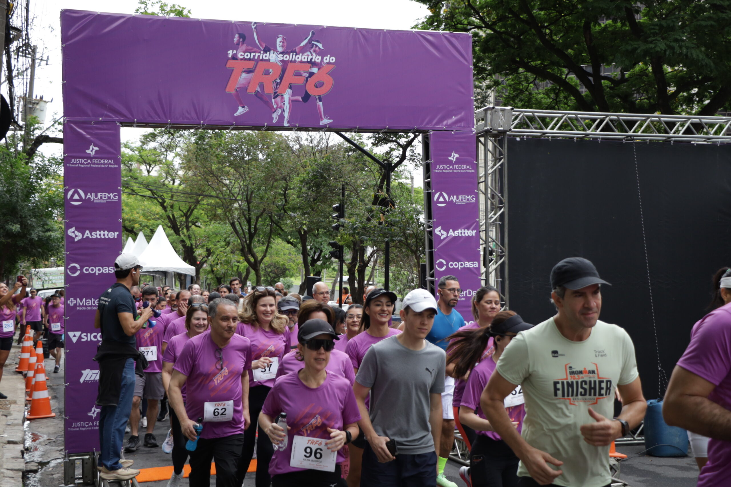 Corredores, a maioria usando camisetas roxas, partem de uma linha de largada sob um grande arco roxo com a inscrição "TRF6" em um evento de corrida de rua, cercado por árvores.