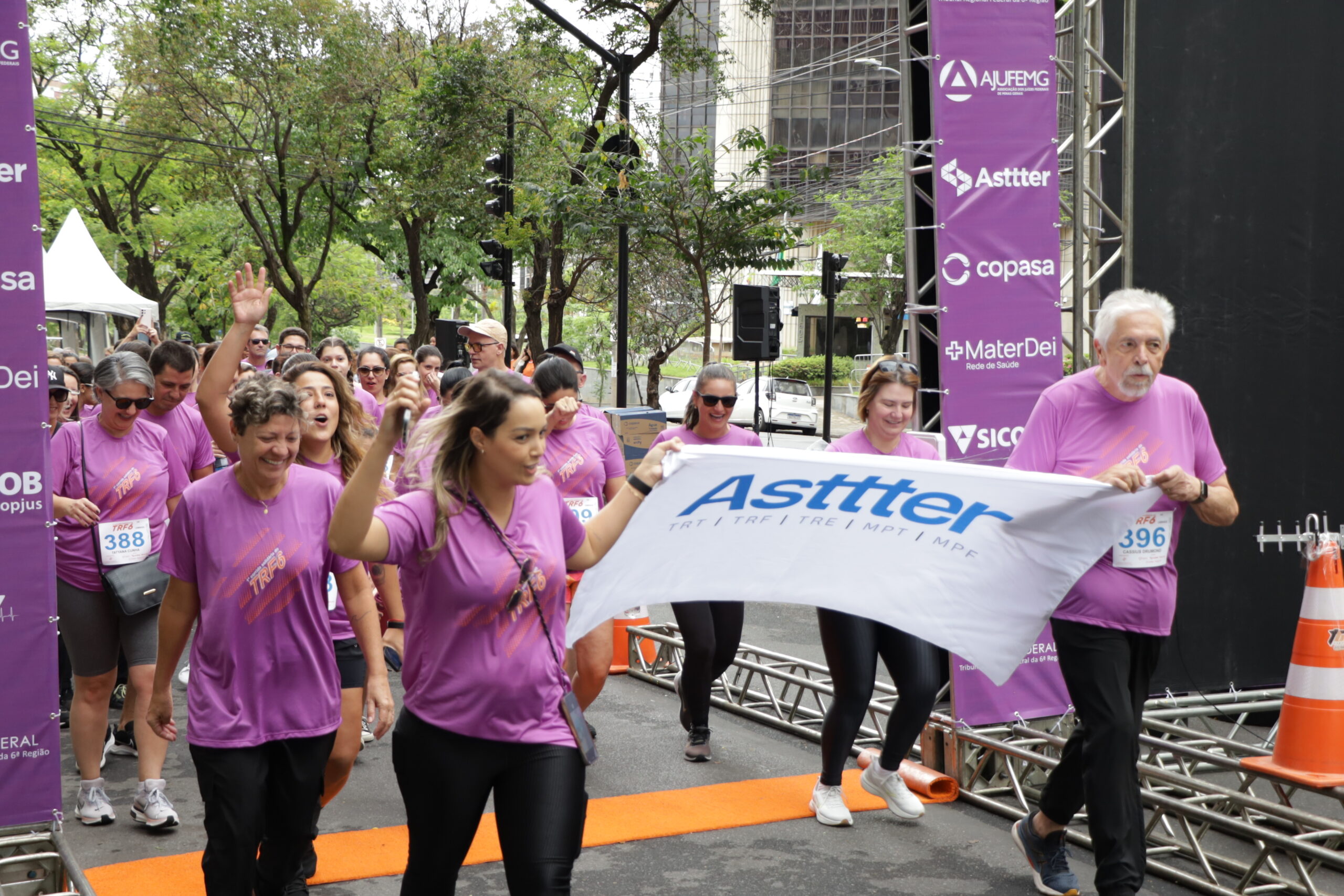 Vários corredores em camisetas roxas de um evento esportivo caminham sob uma estrutura de largada/chegada. Duas pessoas na frente seguram uma faixa branca com o logo "Asttter".