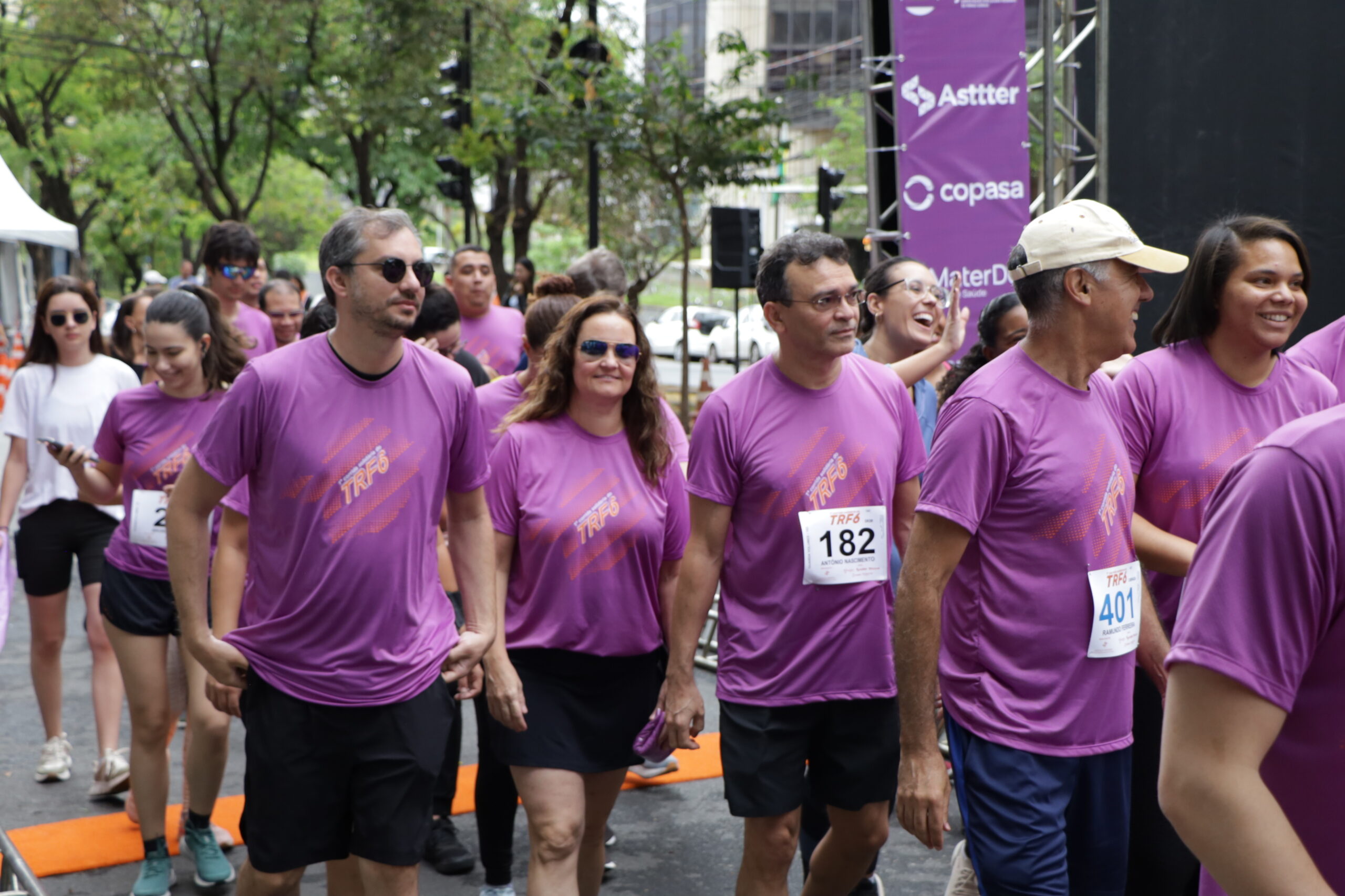 Grupo de corredores, a maioria usando camisetas roxas de um evento esportivo, está caminhando ou correndo em uma rua urbana.