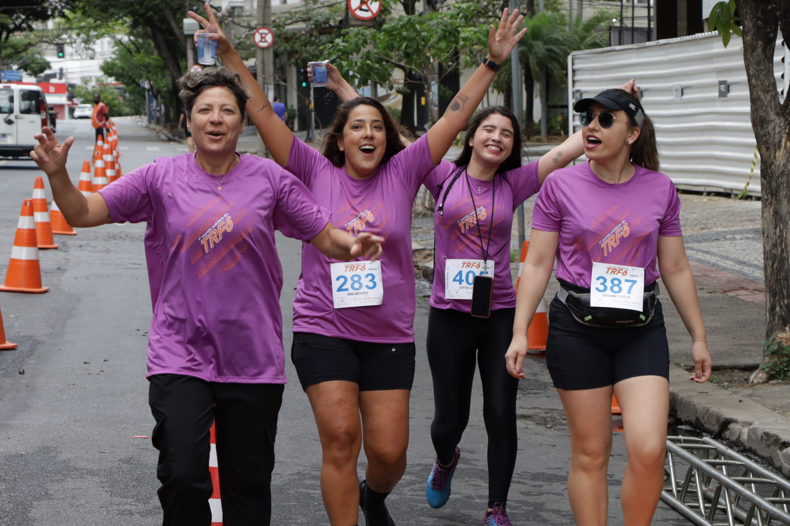 Quatro mulheres em camisetas roxas de corrida sorriem e levantam os braços em celebração enquanto caminham pela rua.