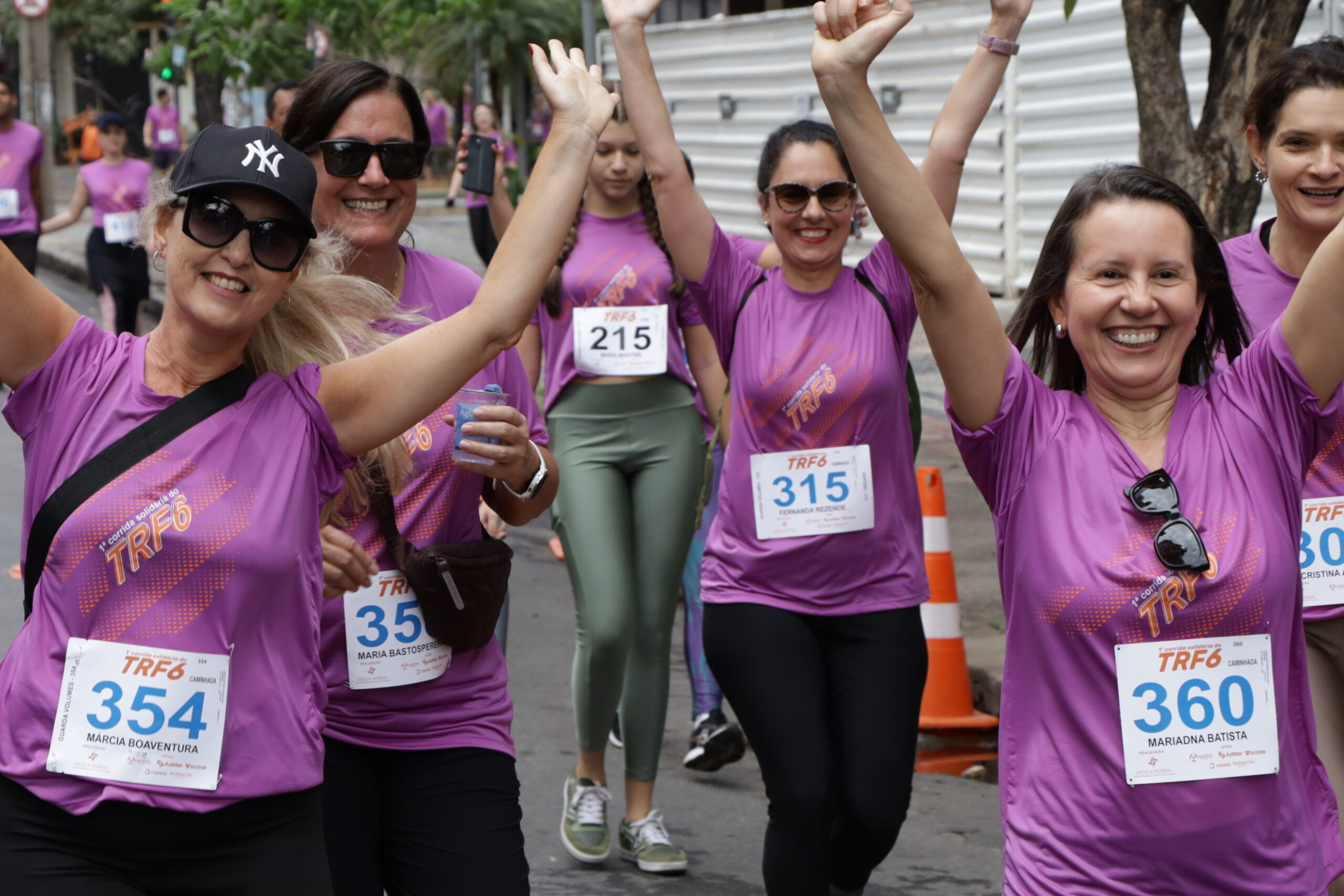 Grupo de mulheres, em camisetas roxas de corrida, sorriem e levantam os braços em celebração enquanto correm na rua.