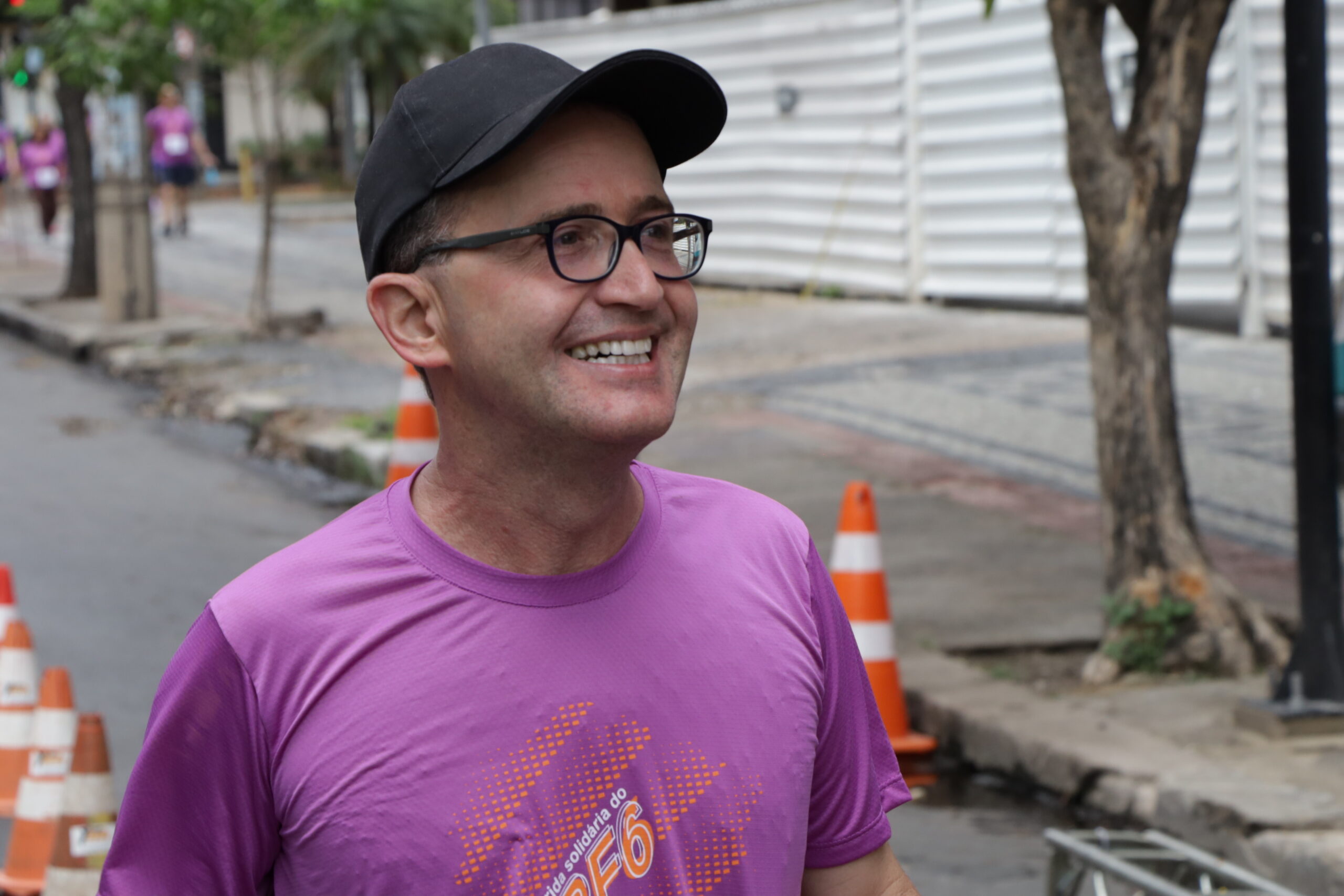 Homem de boné preto e óculos, vestindo uma camiseta roxa de corrida, sorri enquanto caminha na rua.