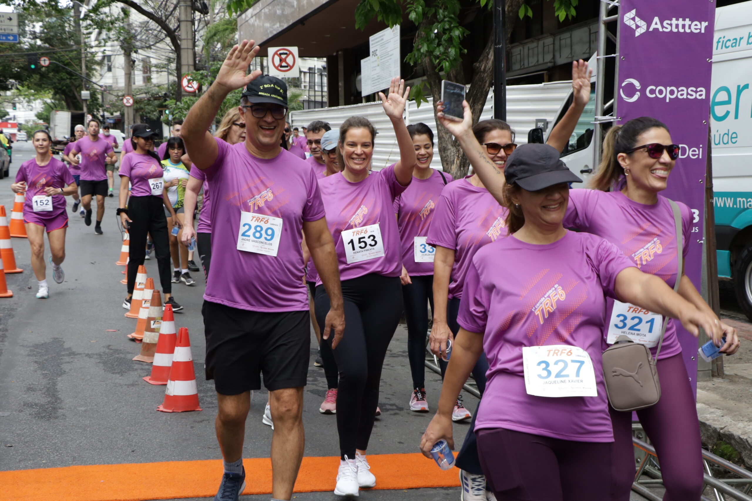 Um grupo de corredores em camisetas roxas, sorrindo e acenando, avança em uma rua urbana durante um evento esportivo.