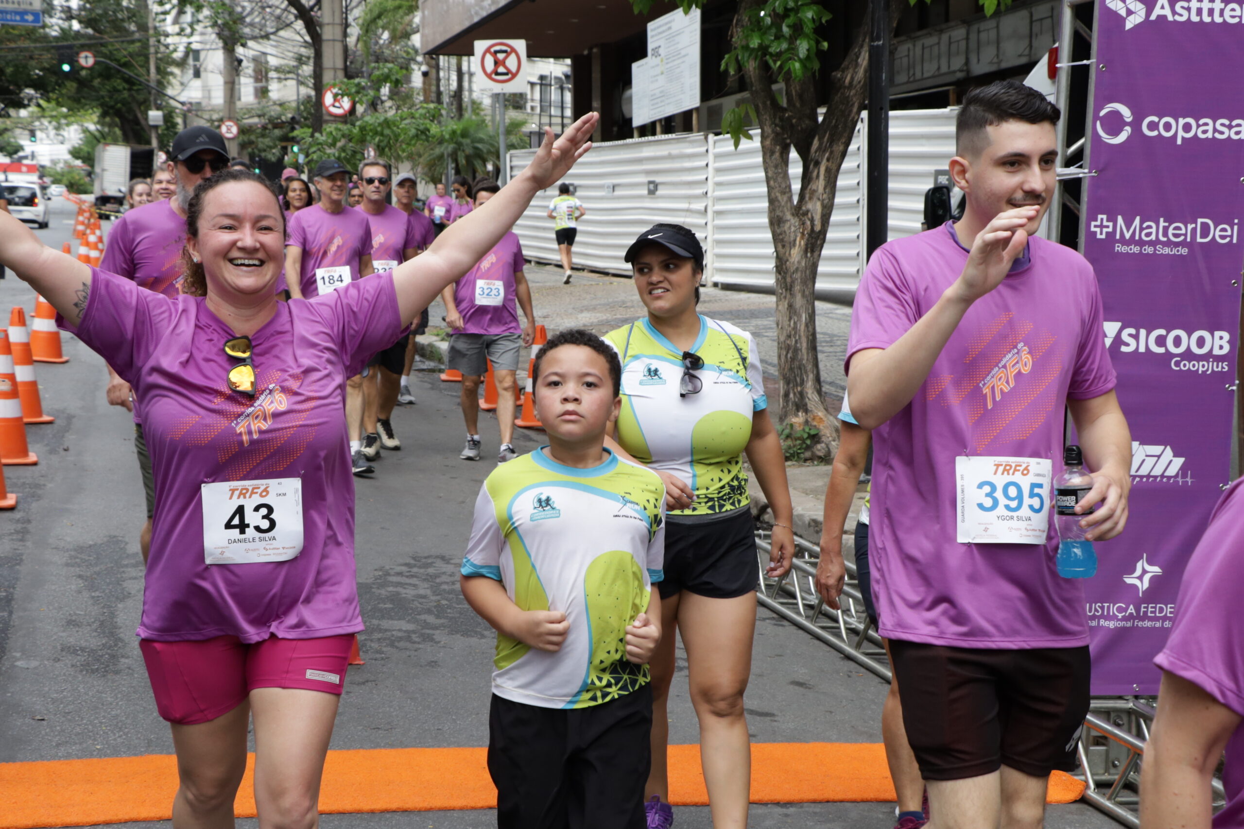 Corredores, a maioria em camisetas roxas, sorriem e celebram enquanto cruzam uma linha de chegada em um evento de rua. Duas pessoas, vestindo uma camiseta verde e branca está no centro.