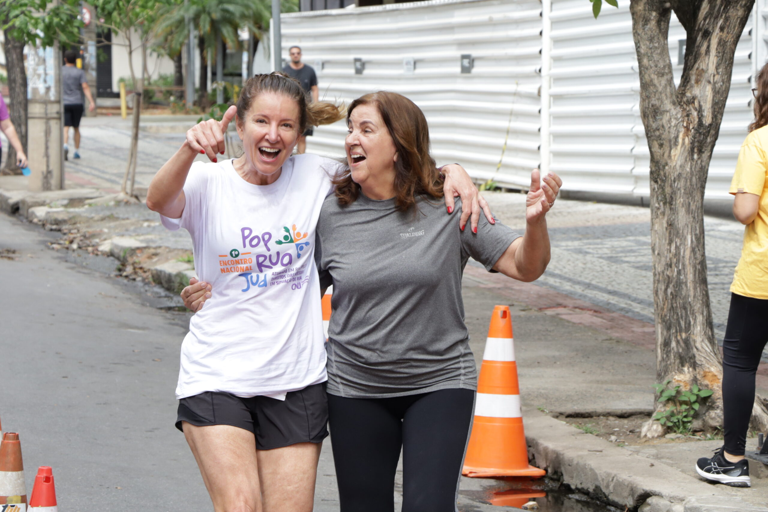 Duas mulheres, lado a lado, sorriem e celebram enquanto correm juntas em uma rua, uma de camiseta branca e outra de camiseta cinza.