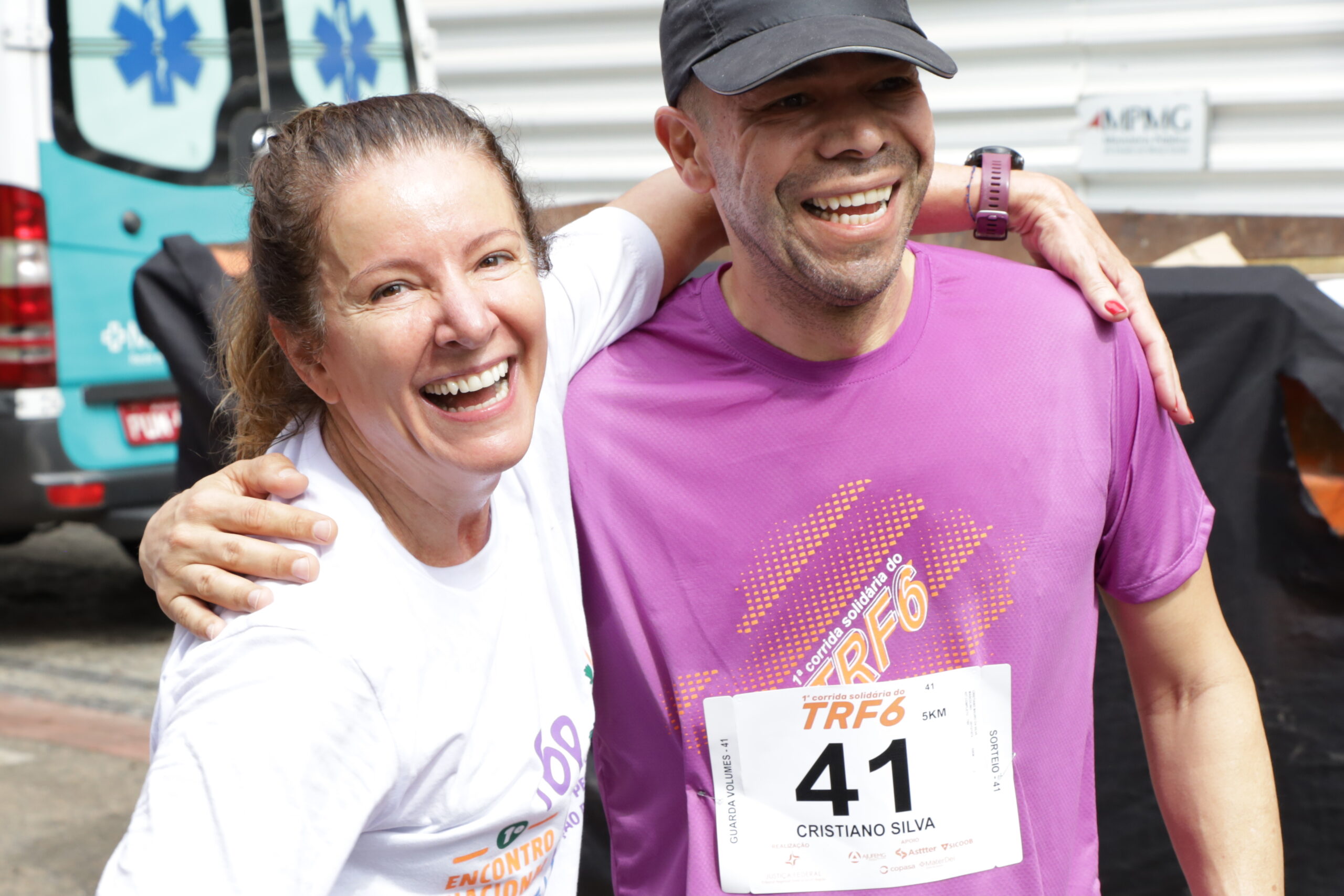 Um homem e uma mulher sorriem e se abraçam após uma corrida. O homem veste uma camiseta roxa e a mulher uma camiseta branca, com uma ambulância no fundo da foto.