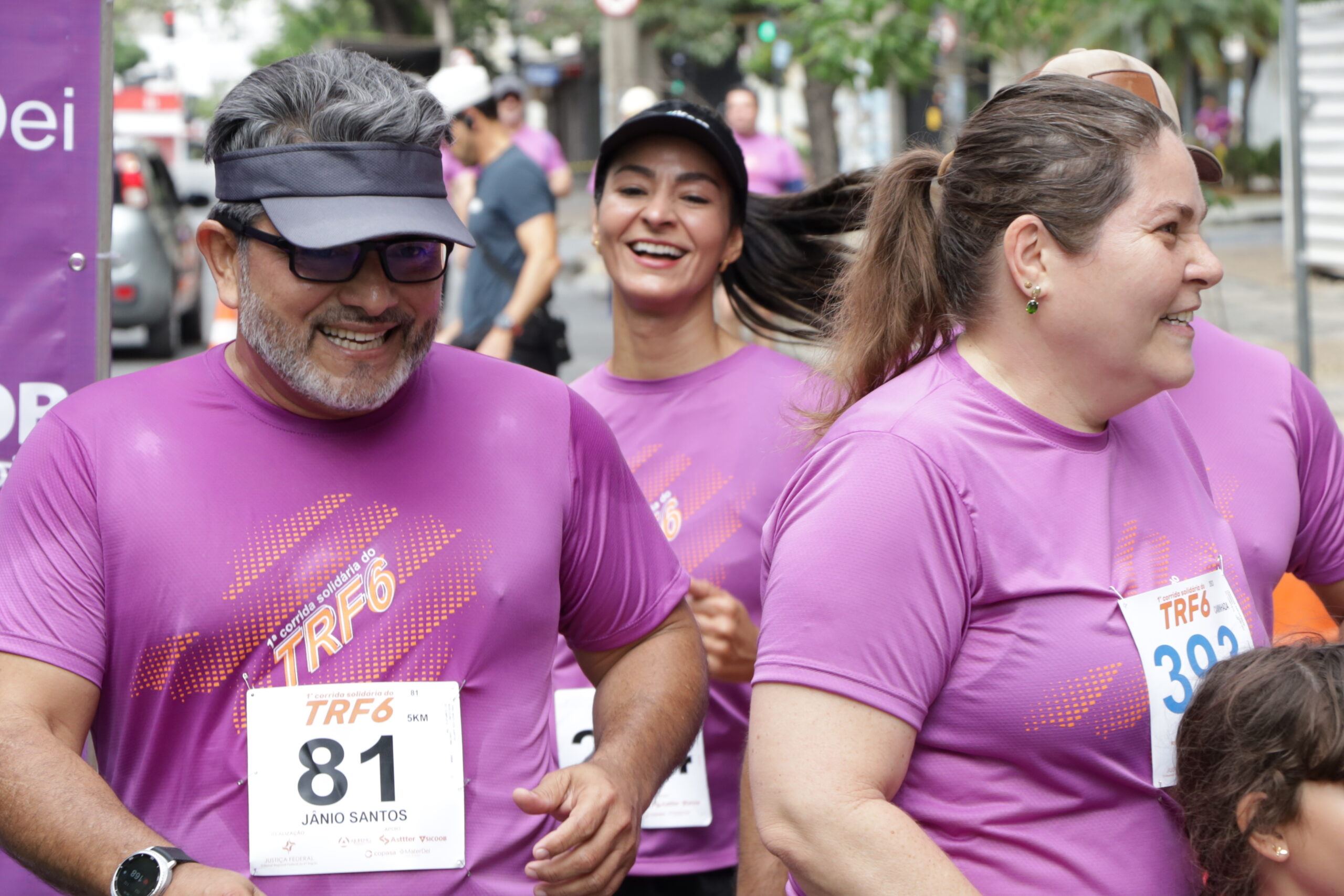 Três corredores sorrindo, vestindo camisetas roxas de um evento com a sigla "TRF6", participam de uma corrida de rua.