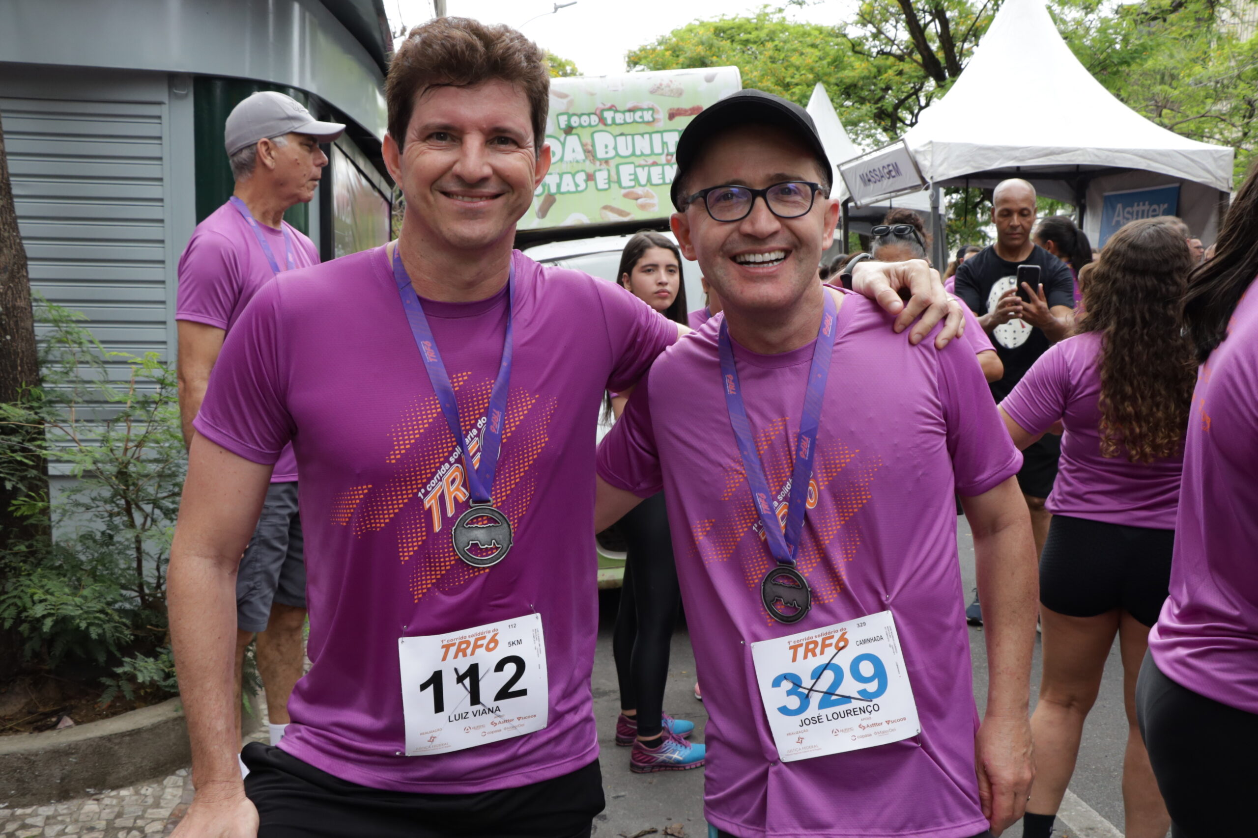 Dois homens, em camisetas roxas de corrida, sorriem e posam abraçados, exibindo medalhas e números de peito, em um evento de rua.