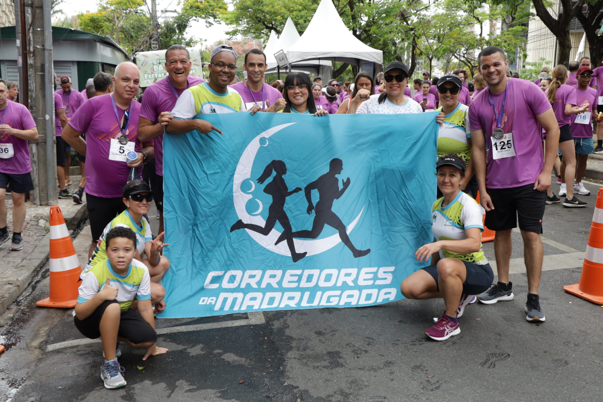 Grupo de corredores sorrindo, em camisetas roxas e brancas, posa segurando uma faixa azul-turquesa com o logo e o texto "CORREDORES DA MADRUGADA" em um evento de rua.