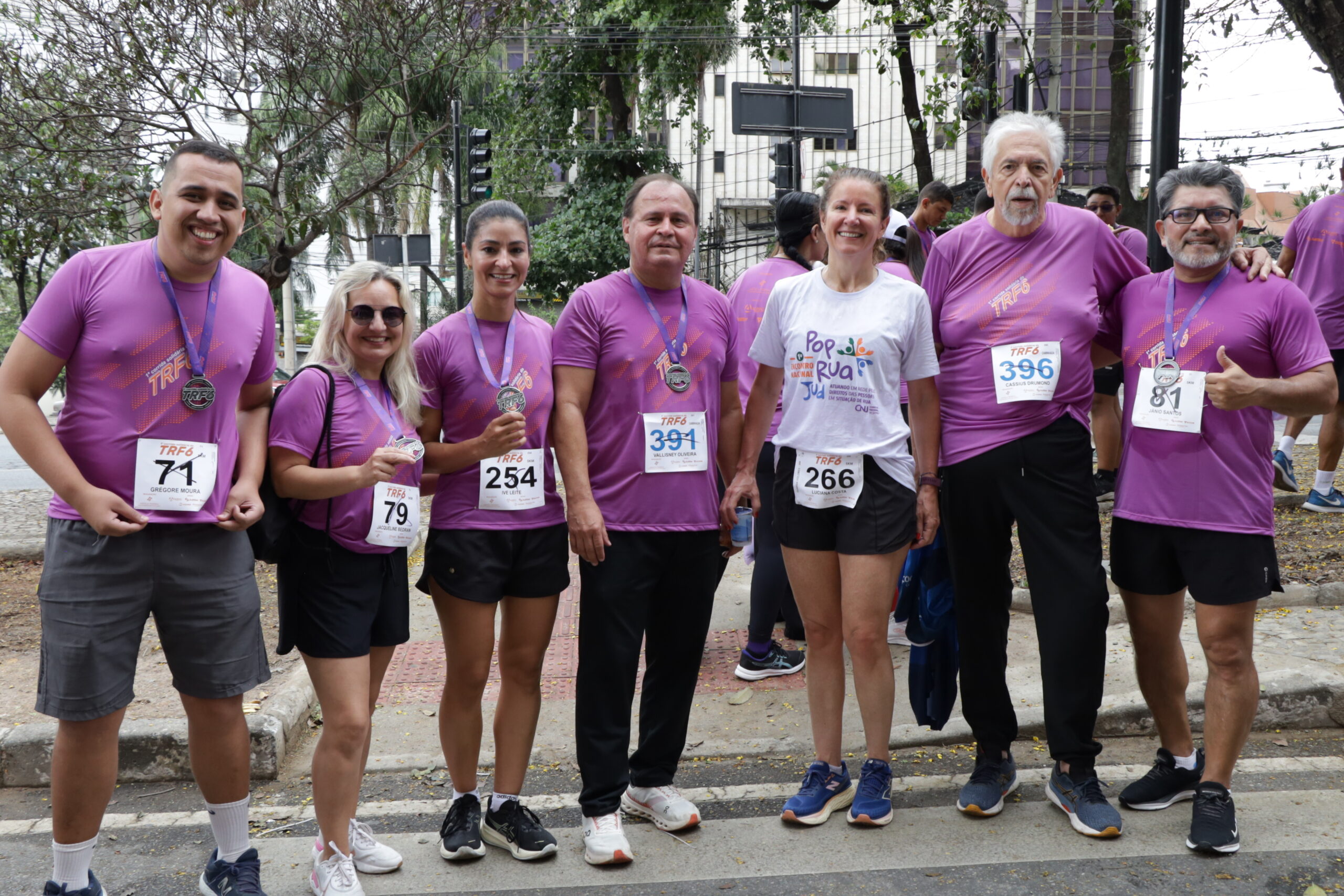 Sete corredores (quatro homens e três mulheres) em camisetas roxas (exceto uma mulher de camiseta branca), sorriem e posam com suas medalhas e números de peito após a corrida.