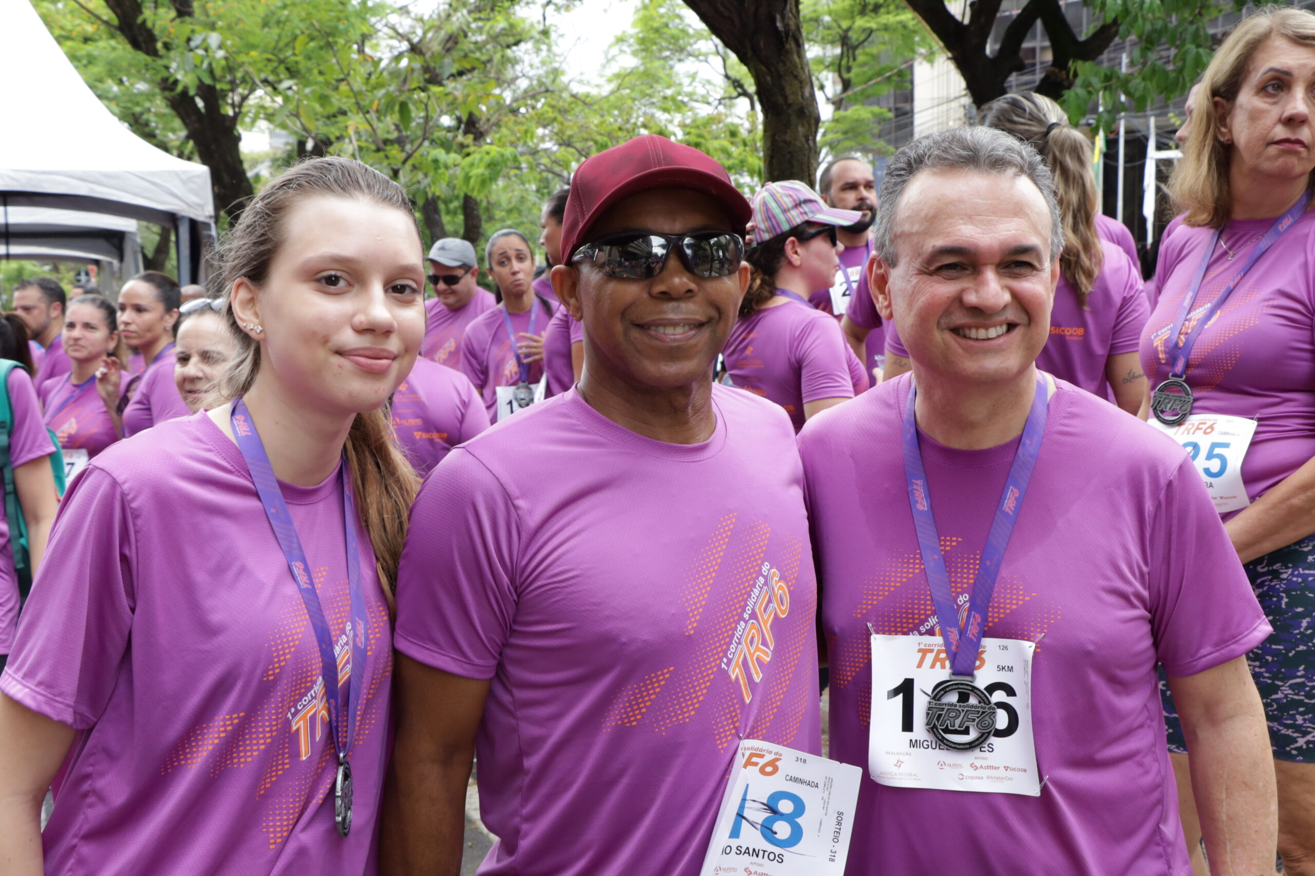 Três corredores em camisetas roxas, com medalhas no pescoço, sorriem para a câmera em meio a uma multidão após o evento.