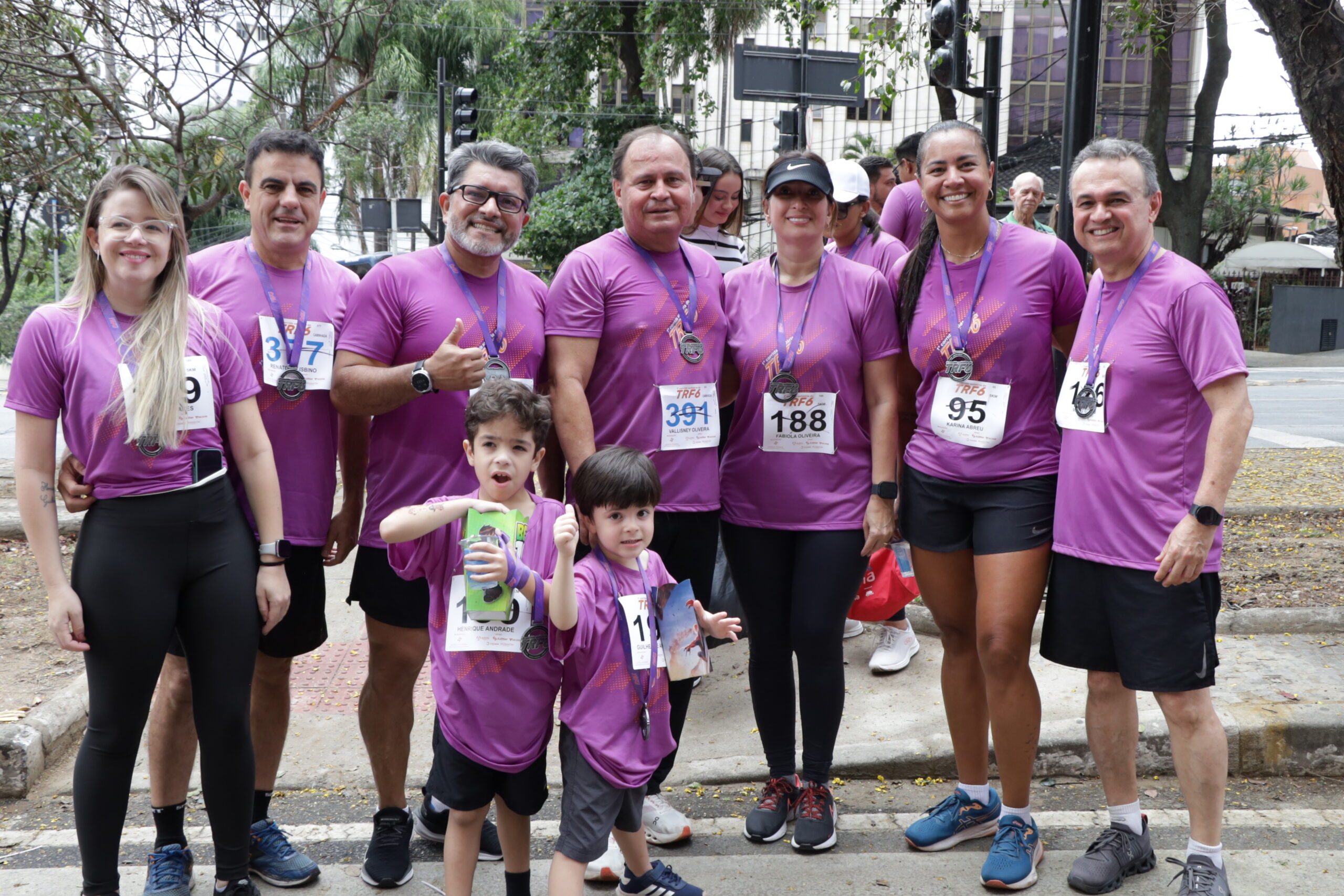 Grande grupo de adultos e duas crianças, todos vestindo camisetas roxas de corrida e a maioria com medalhas, sorriem e posam juntos ao ar livre.