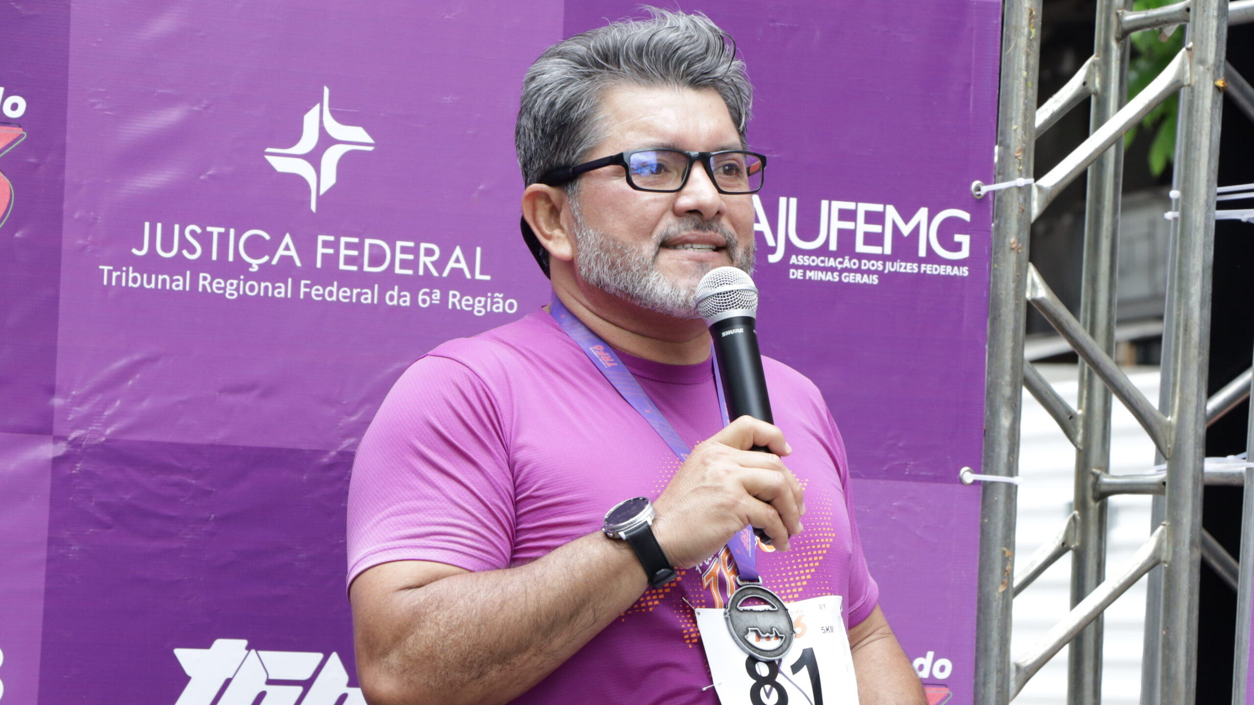 Homem de barba grisalha, óculos, camiseta roxa de corrida e medalha, fala ao microfone em frente a um banner roxo com logos da Justiça Federal TRF6 e da AJUFEMG.