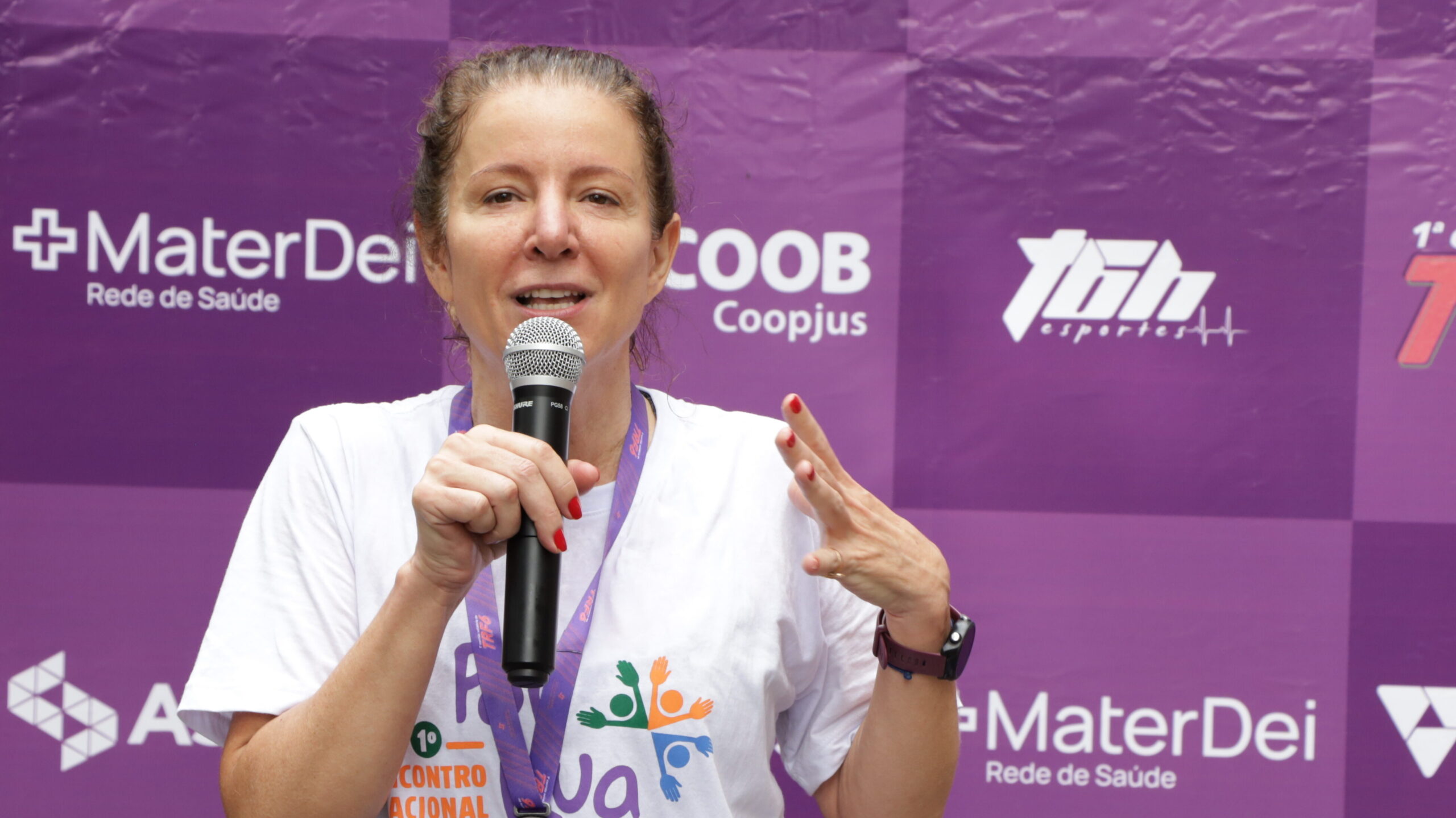 Mulher de camiseta branca fala ao microfone, gesticulando com a mão, em frente a um banner roxo com logos de patrocinadores.
