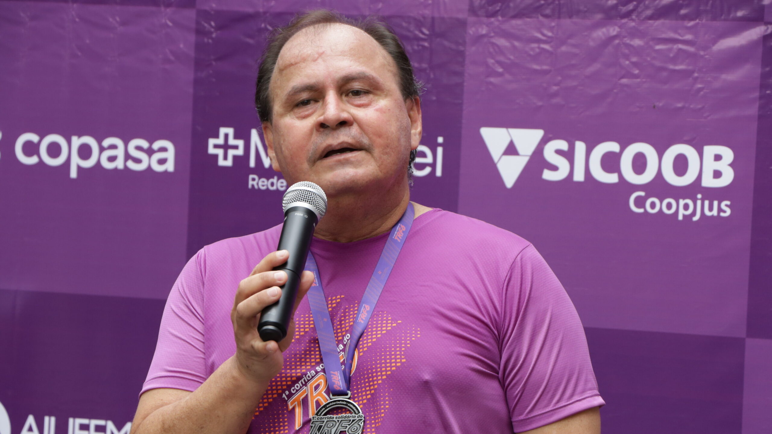Homem de camiseta roxa, com medalha de corrida no pescoço, fala em um microfone em frente a um banner roxo com logos de patrocinadores.