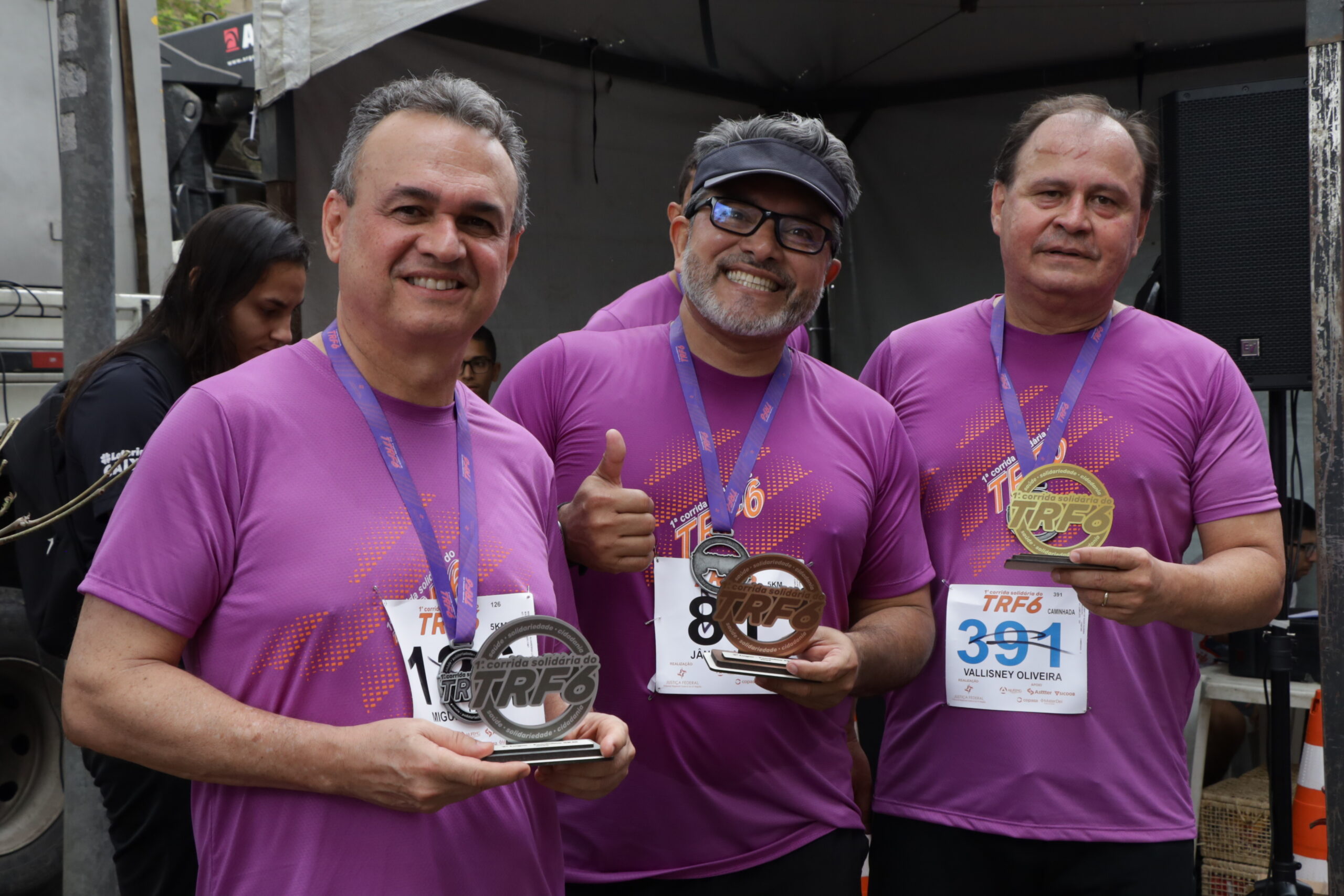 Três homens, em camisetas roxas de corrida, sorriem e exibem suas medalhas, em frente a uma barraca.