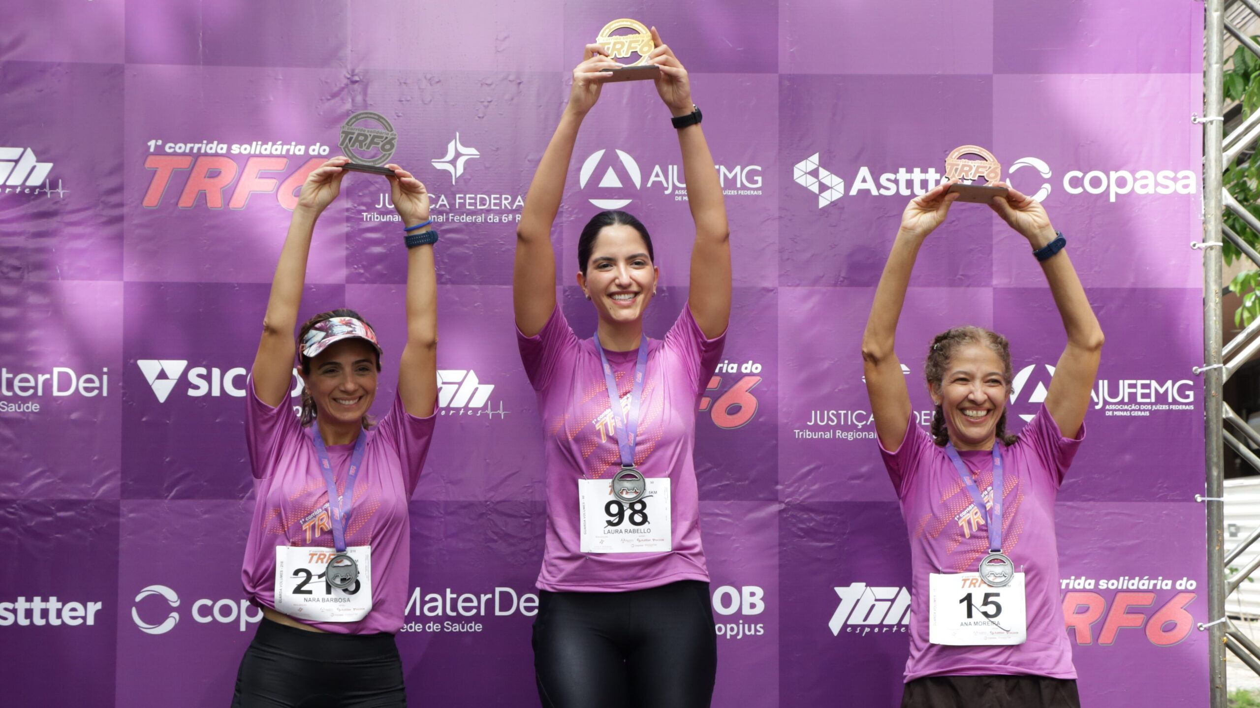 Três mulheres, usando camisetas roxas, medalhas e números de peito, celebram no pódio, segurando seus troféus acima da cabeça, em frente a um banner roxo de patrocinadores.