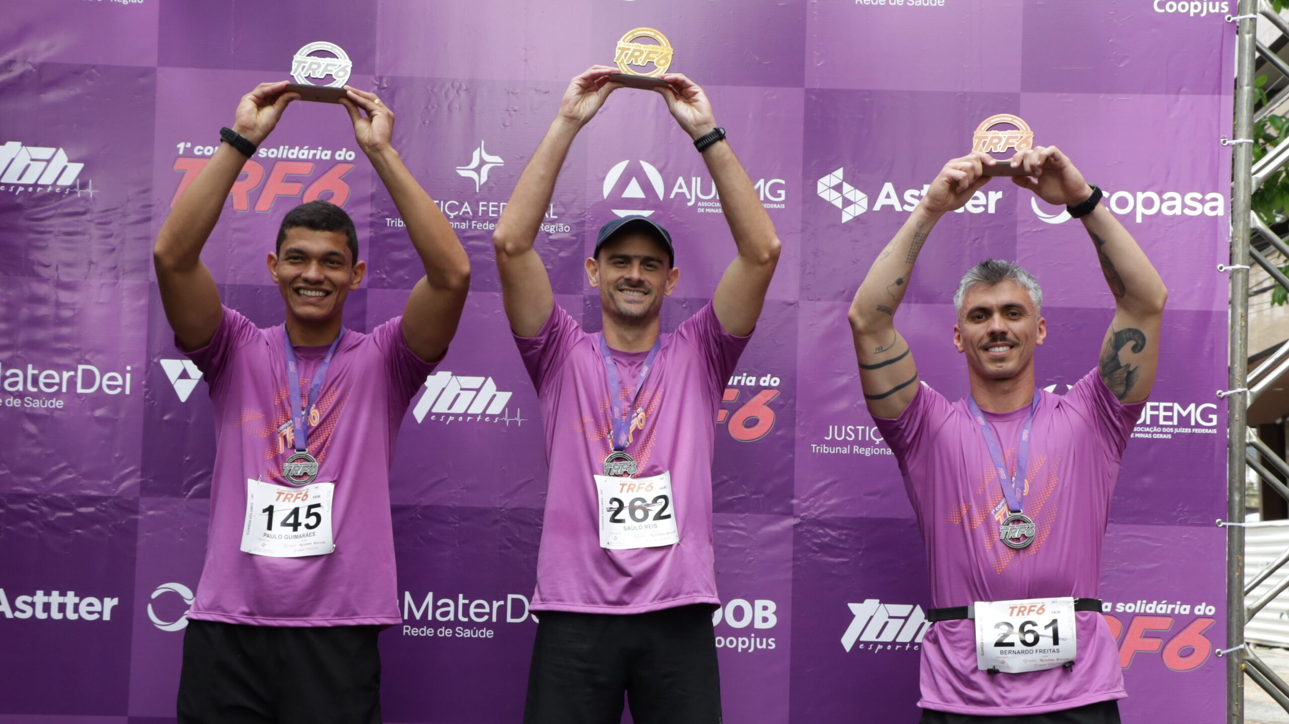 Três homens, em camisetas roxas de corrida e medalhas, celebram no pódio, segurando seus troféus acima da cabeça, em frente a um banner roxo de patrocinadores.