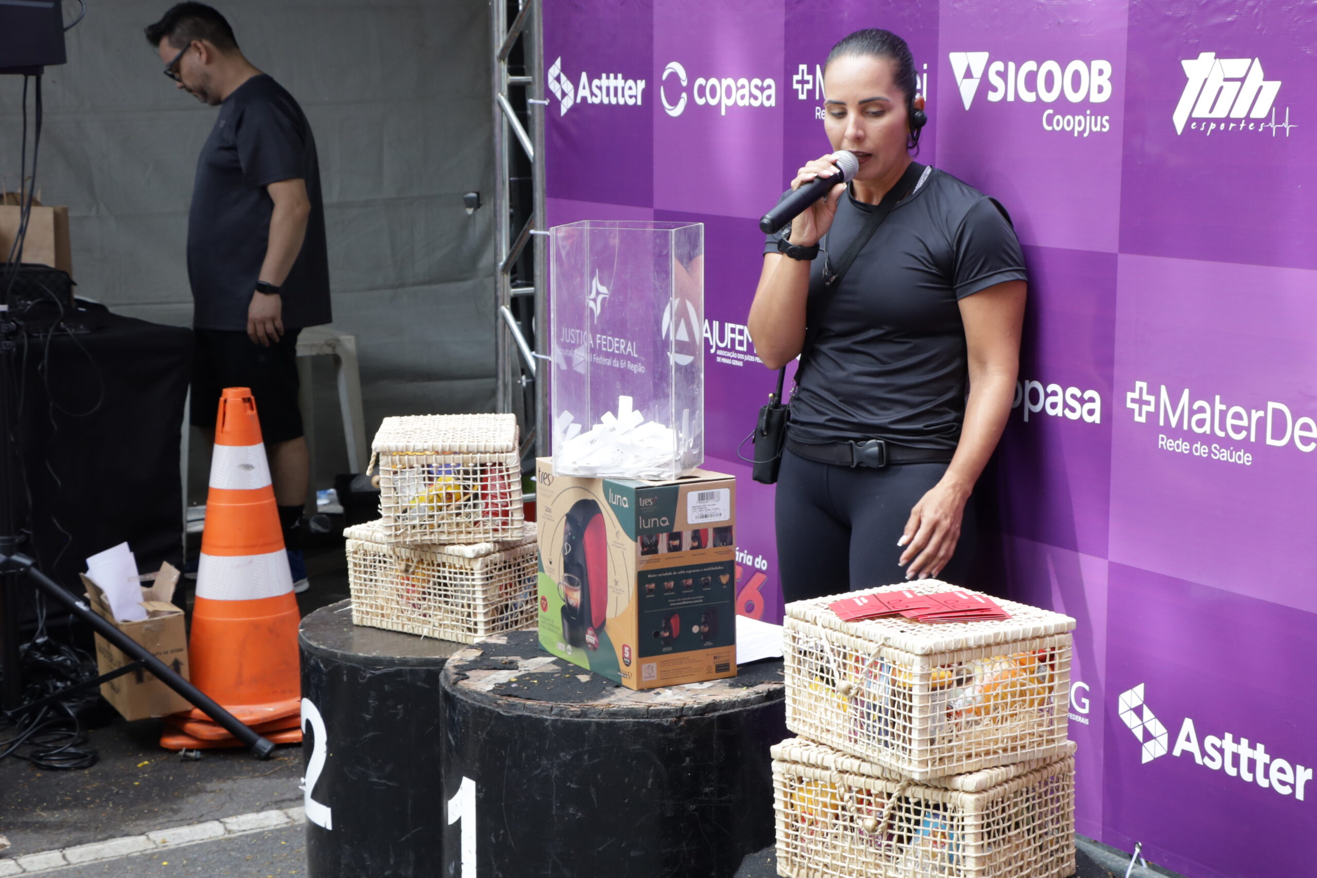 Mulher de roupa preto fala ao microfone ao lado de caixas de sorteio em um palco com fundo roxo de patrocinadores durante um evento.