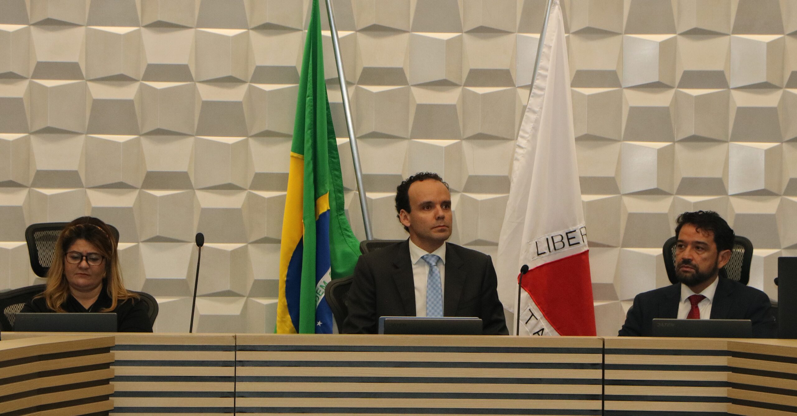 Três pessoas (uma mulher e dois homens) vestindo trajes formais estão sentadas em uma mesa em um ambiente institucional, com as bandeiras do Brasil e de Minas Gerais ao fundo.