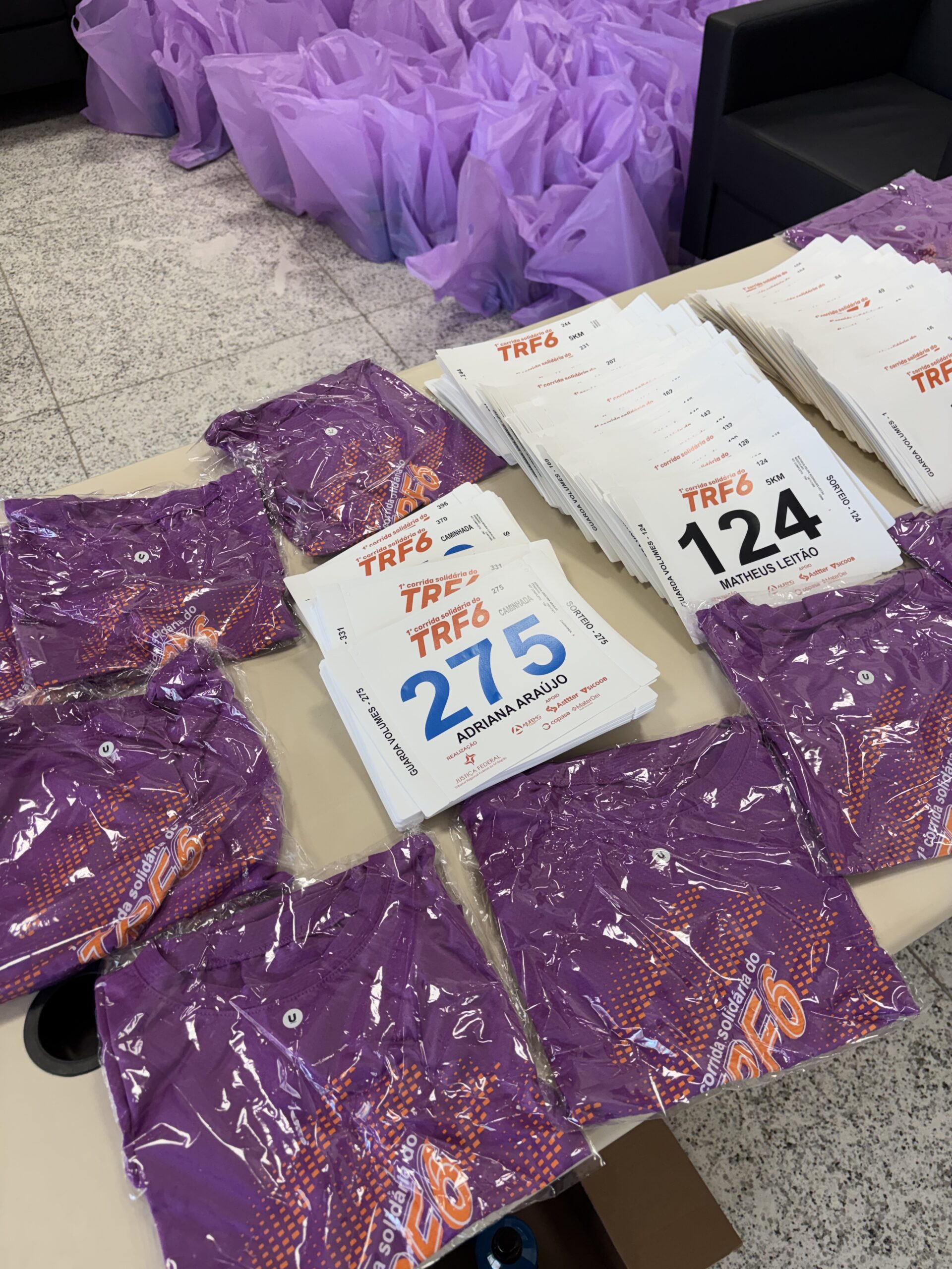 A imagem mostra uma mesa com materiais prontos para distribuição. Sobre a mesa, estão dispostas várias camisetas roxas, embaladas individualmente em plástico transparente, com uma estampa laranja do "TRF6" visível. Ao lado e empilhados, há vários números de peito brancos para corrida, com a identificação "TRF6" no topo e grandes números e nomes de corredores. Ao fundo, no chão ou em outra superfície, há uma grande quantidade de sacolas plásticas roxas amassadas, possivelmente contendo os kits completos do evento.