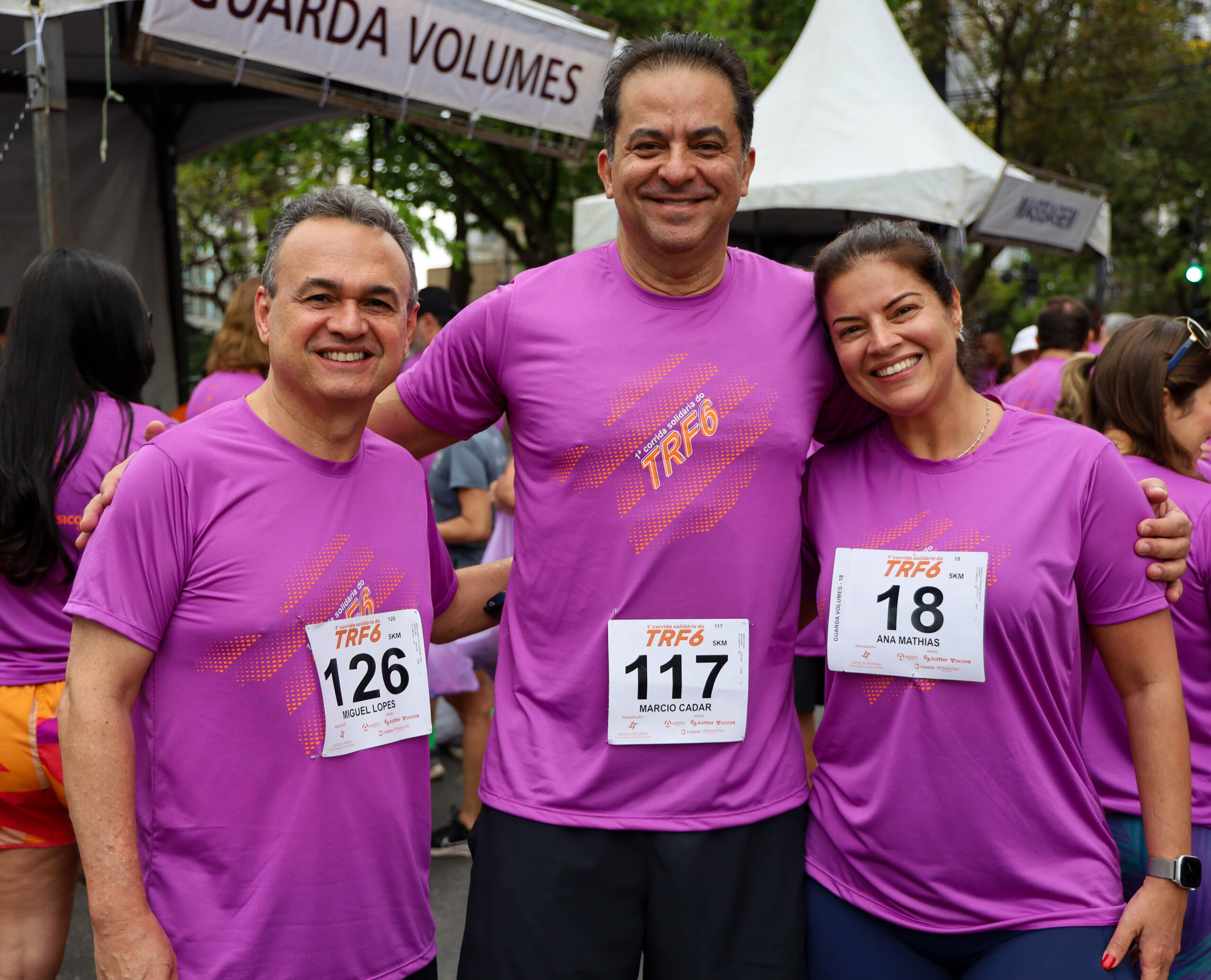 Três corredores (dois homens e uma mulher) em camisetas roxas de corrida e números de peito, sorriem abraçados em um evento esportivo.