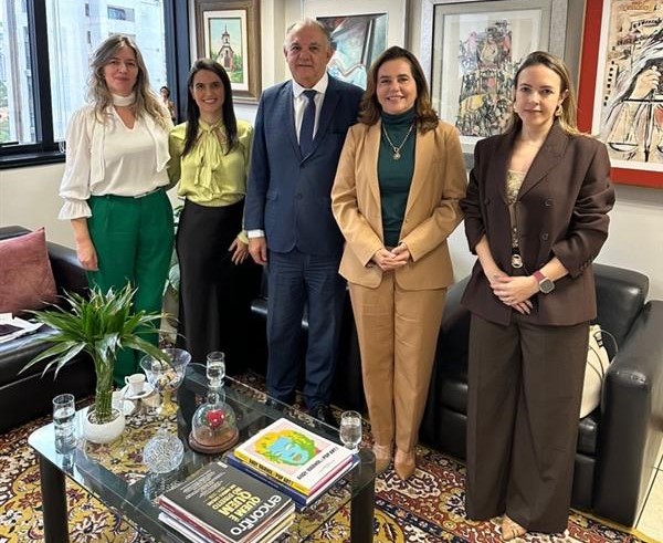 Uma fotografia de grupo mostra cinco pessoas (um homem e quatro mulheres) posando em pé em um escritório. O homem veste um terno. As mulheres vestem trajes sociais, incluindo calças sociais, blazers e blusas. Em primeiro plano, há uma mesa de centro com livros e copos, e obras de arte estão penduradas nas paredes do fundo.