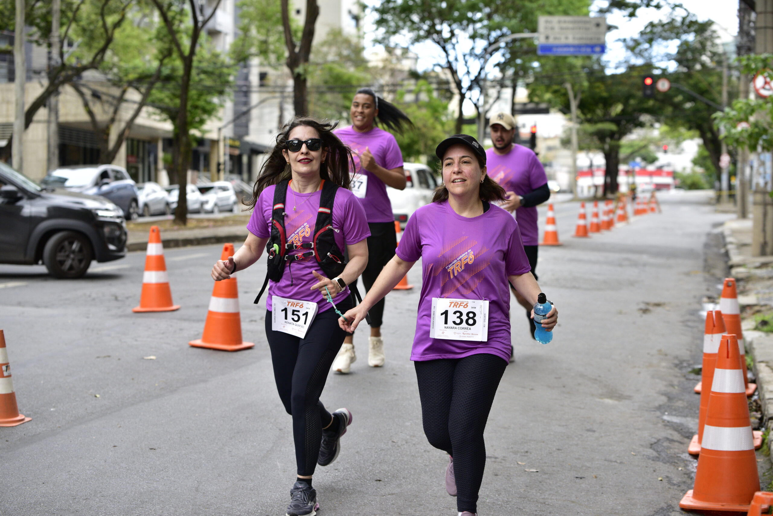 Quatro corredores em camisetas roxas, com números de peito, correm em uma rua urbana delimitada por cones de trânsito.