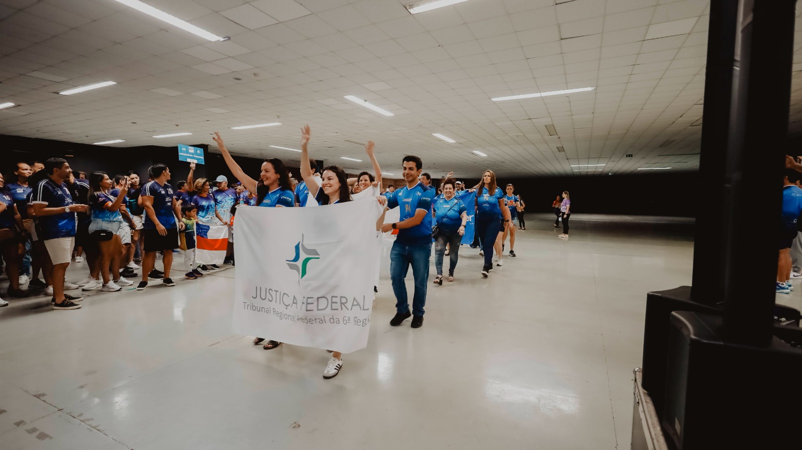 Foto em um ginásio ou grande salão coberto, mostrando um grupo de pessoas em marcha. Duas mulheres na frente seguram uma bandeira branca com o logotipo e texto "JUSTIÇA FEDERAL Tribunal Regional Federal da 6ª Região". O grupo está vestido com camisetas azuis (uniformes), muitas pessoas erguem os braços em celebração.