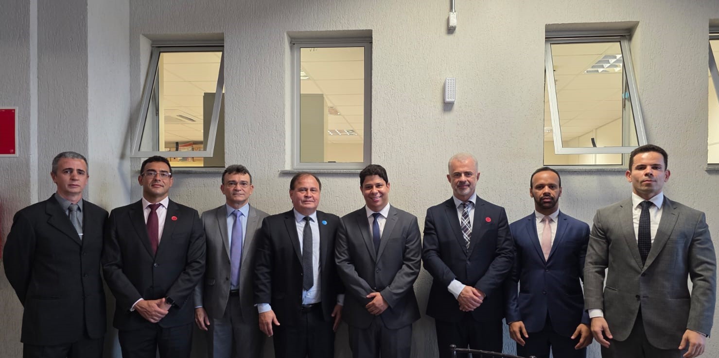 A imagem é uma fotografia de grupo de oito homens, que estão em pé e alinhados em um ambiente interno. Todos os homens estão vestindo ternos e gravatas em tons variados de cinza e preto. Eles estão posicionados em frente a uma parede de cor clara que apresenta uma fileira de três janelas retangulares abertas na parte superior.