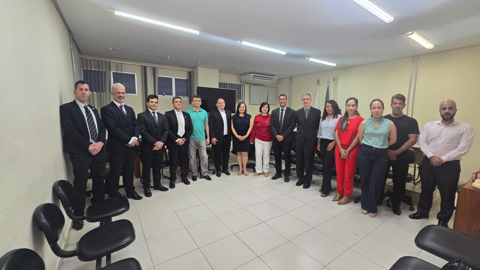 A imagem é uma fotografia de um grupo de pessoas, que estão em pé e alinhadas em semicírculo em um ambiente interno que parece ser um escritório ou sala de reunião. O grupo é composto por homens e mulheres. Muitos dos homens estão vestindo ternos e gravatas, enquanto outros usam camisas sociais ou roupas casuais. As mulheres vestem roupas sociais ou blusas de cores vivas e calças ou saias. O ambiente possui paredes claras, piso de cerâmica clara, iluminação de teto e cadeiras de escritório escuras em primeiro plano e nas laterais.