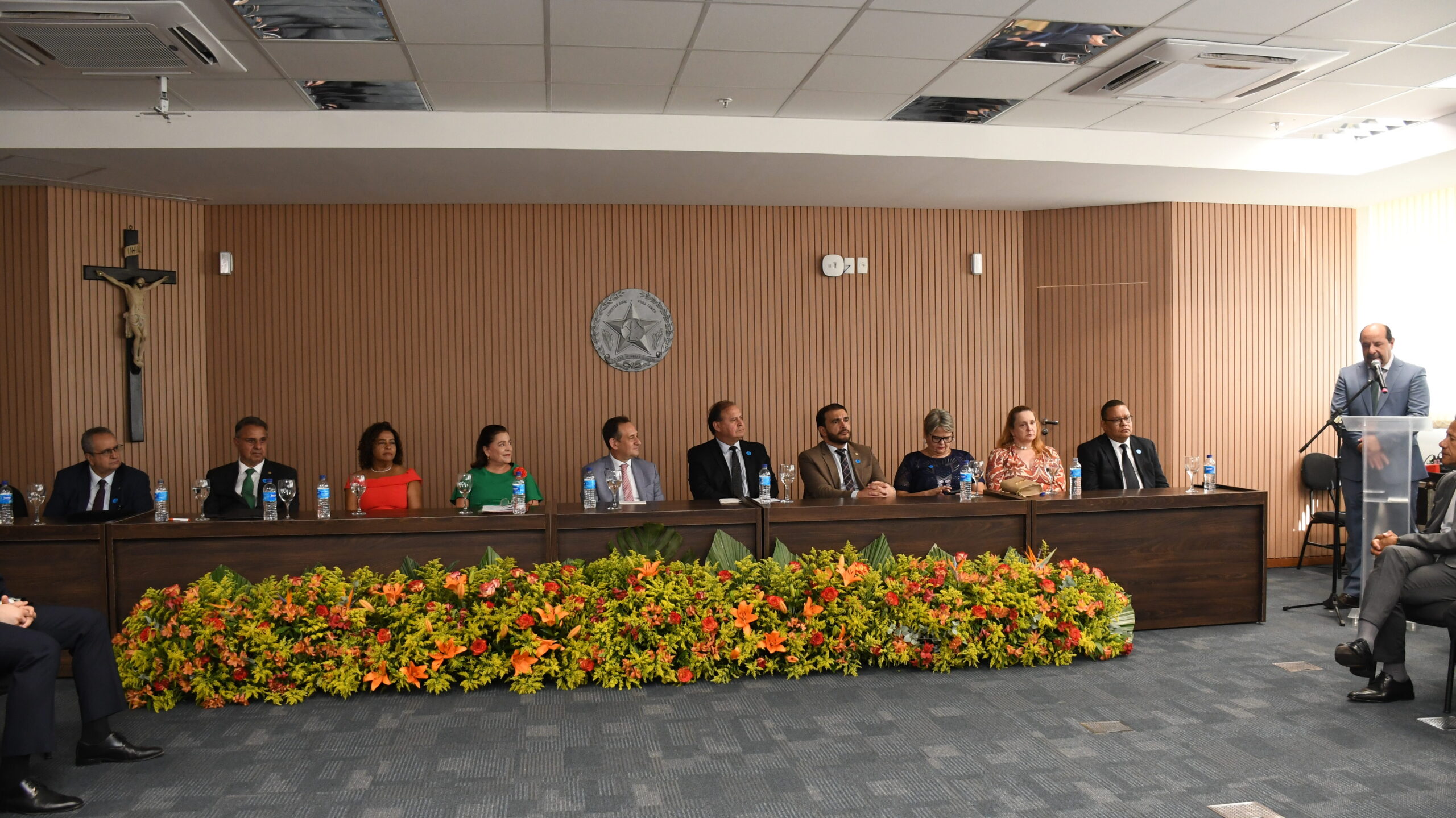 A imagem mostra um ambiente formal interno, com uma longa mesa de conferência atrás de um arranjo floral grande em tons de amarelo, laranja e verde. Dez pessoas, homens e mulheres, estão sentadas à mesa, todas vestindo trajes sociais (ternos, blazers ou vestidos coloridos). Em frente à mesa, à direita, um homem de terno está em pé em um púlpito, falando. Ao fundo, a parede de madeira ripada possui um crucifixo à esquerda e um brasão da República ou emblema institucional no centro. O chão é forrado com carpete escuro.