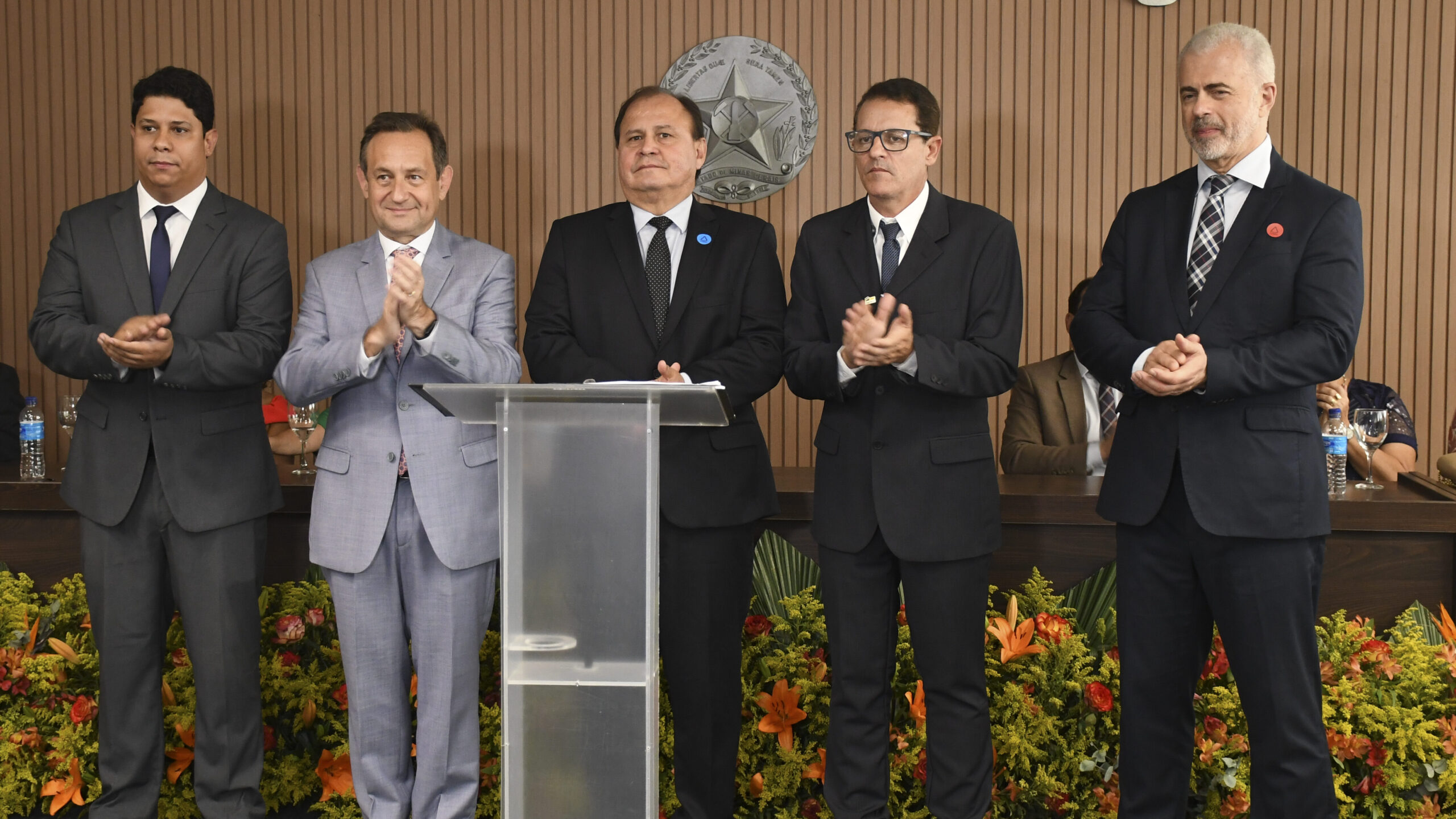 A imagem é uma fotografia que mostra cinco homens em pé em um palco ou plataforma durante uma cerimônia formal. Todos os homens estão vestidos com ternos e gravatas, sendo a maioria em tons escuros, exceto o segundo da esquerda, que veste um terno cinza claro. Eles estão atrás de um púlpito de acrílico e parecem estar aplaudindo. A decoração do palco inclui arranjos florais em tons de laranja e verde na frente. Ao fundo, uma parede de madeira escura tem um grande emblema circular fixado.