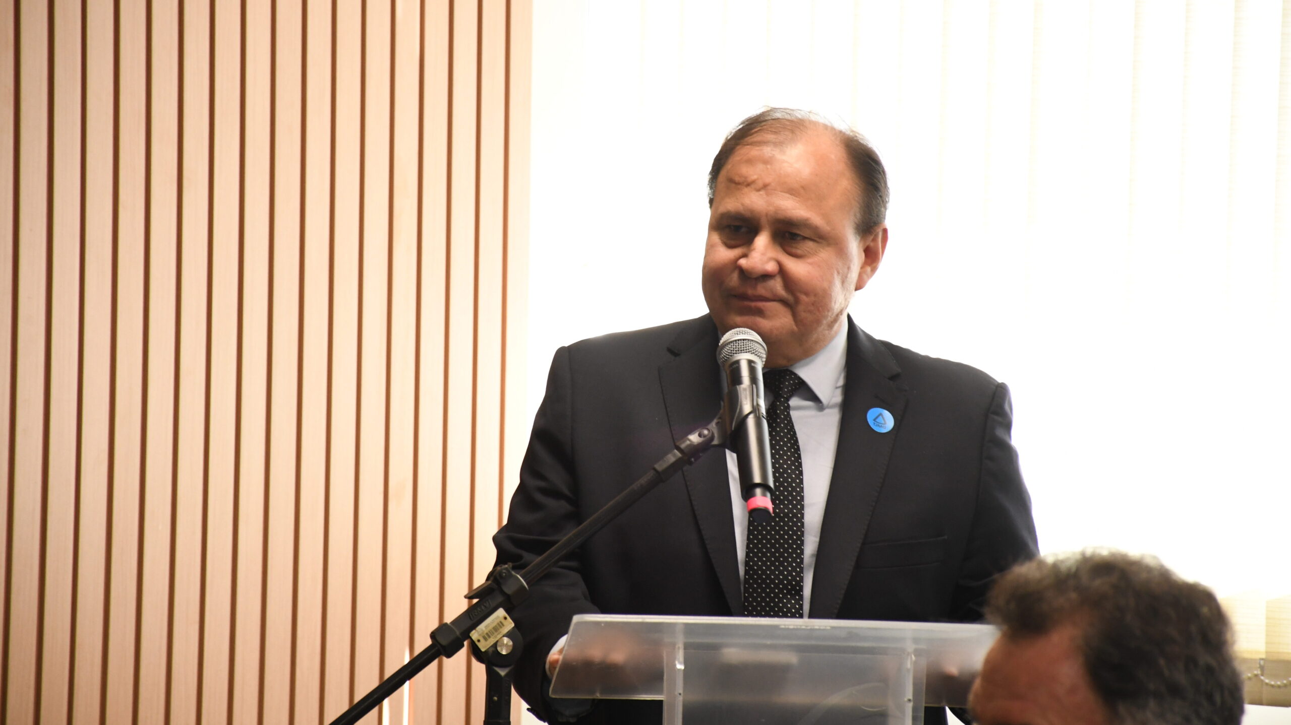A imagem é um close-up de um homem de meia-idade, vestido com um terno escuro e gravata de bolinhas, em pé atrás de um púlpito transparente, com um microfone à sua frente. Ele está olhando para a direita. O fundo é composto por uma parede de madeira ripada à esquerda e uma área de luz clara.