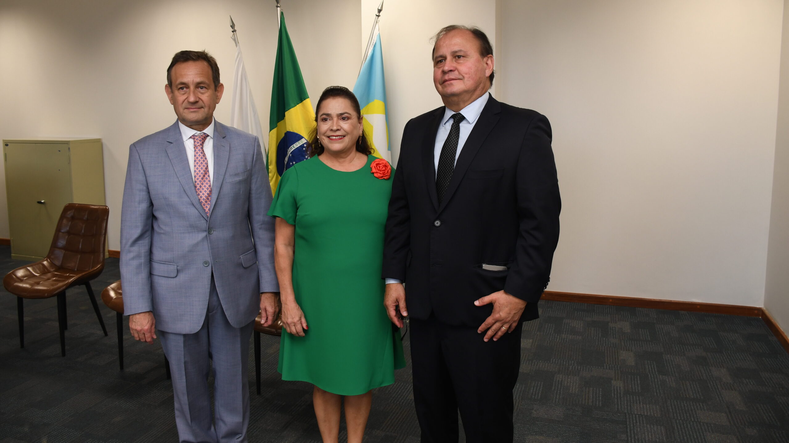 A imagem é uma fotografia de três pessoas (dois homens e uma mulher) posando juntas em um ambiente interno. O homem à esquerda veste um terno cinza claro e gravata estampada. A mulher ao centro veste um vestido verde com um broche de flor vermelha e o outro homem usa terno escuro. Atrás deles, há três bandeiras hasteadas. O ambiente tem paredes claras e um carpete escuro, com alguns móveis visíveis à esquerda.
