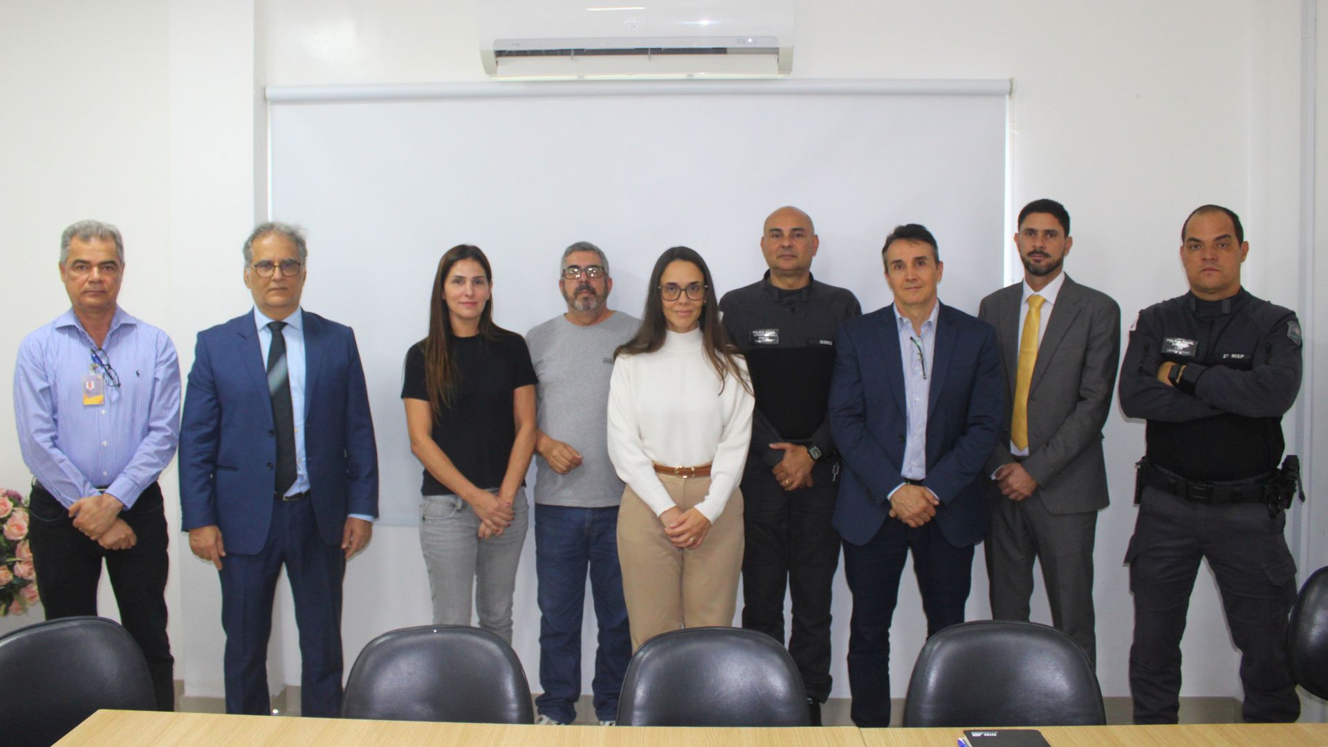 A imagem é uma fotografia de grupo de nove pessoas — sete homens e duas mulheres — posando em fila em um ambiente interno simples. O grupo exibe uma variedade de vestimentas, incluindo dois homens de terno e gravata, um homem de uniforme tático escuro, e outros de camisas sociais casuais. A mulher à frente, no centro, veste uma blusa de gola alta clara e calça social. Ao fundo, uma parede branca lisa com uma persiana. Em primeiro plano, há cadeiras de escritório escuras e a borda de uma mesa.