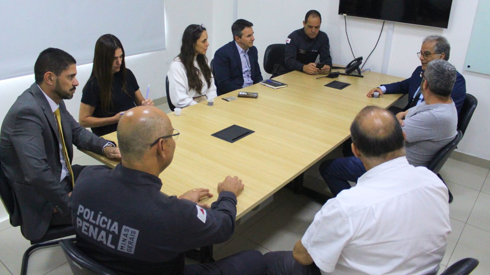 A imagem mostra um grupo de nove pessoas reunidas ao redor de uma mesa de reunião retangular clara, em um ambiente de escritório. Os participantes estão vestidos com trajes formais e casuais variados. Todos estão sentados, e a mesa contém poucos objetos, como cadernos e telefones.