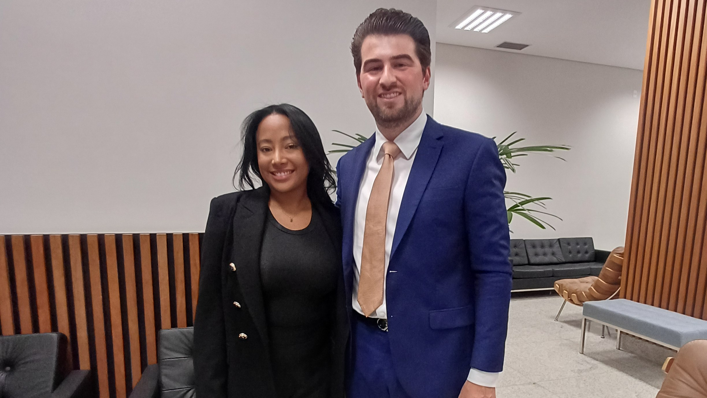 Uma mulher de cabelos pretos e um homem de barba, ambos sorridentes, posam lado a lado em um ambiente moderno com sofás de couro e painéis de madeira ao fundo. Ela veste um casaco preto sobre uma blusa da mesma cor, enquanto ele usa um terno azul-marinho com camisa branca e gravata bege.