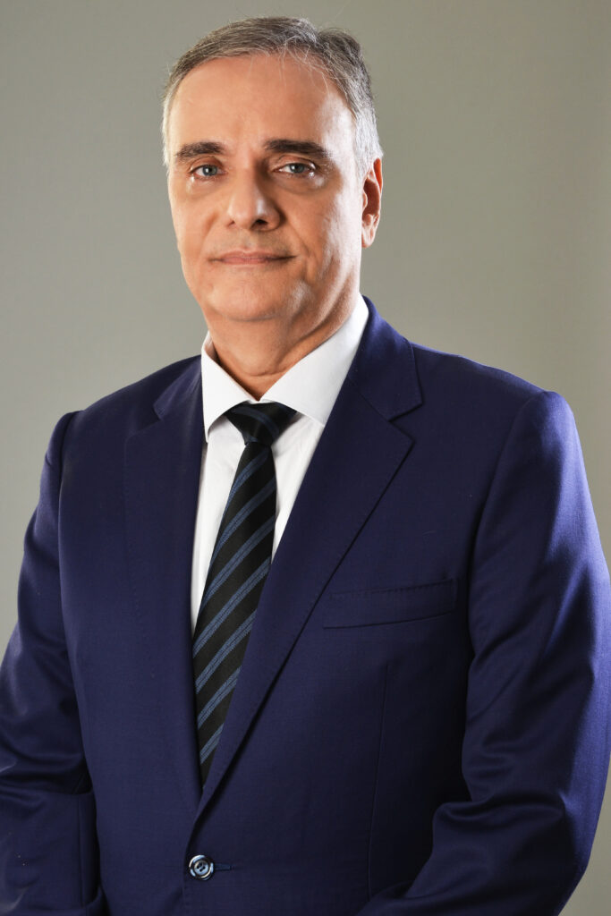 Retrato formal de um homem de meia-idade com cabelos grisalhos curtos, vestindo um terno azul-marinho, camisa branca e gravata escura com listras diagonais sutis. Ele olha diretamente para a câmera com uma expressão séria e composta contra um fundo cinza neutro.