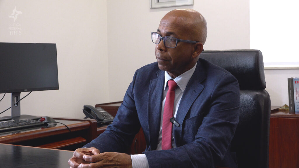 Um homem de óculos e terno azul-marinho com gravata vermelha está sentado em um escritório, com as mãos entrelaçadas sobre a mesa. Ele olha para o lado com expressão séria, posicionado à frente de uma estação de trabalho com monitor, teclado e telefone.