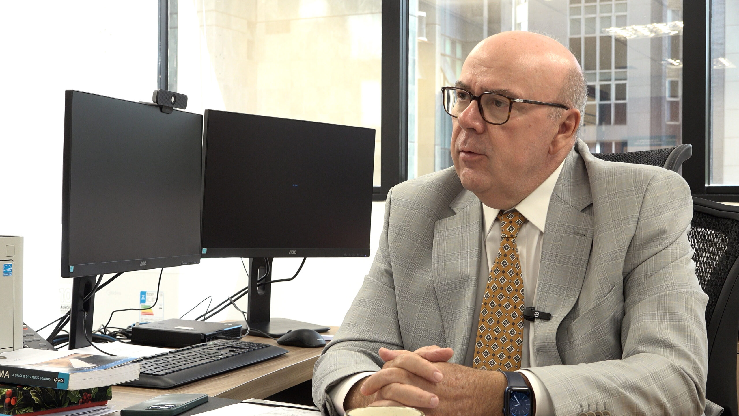 Um homem careca e de óculos, vestindo um terno cinza xadrez com gravata amarela estampada, está sentado à frente de dois monitores de computador em um escritório. Ele mantém as mãos entrelaçadas sobre a mesa e olha atentamente para o lado, enquanto um microfone de lapela está preso à sua lapela.