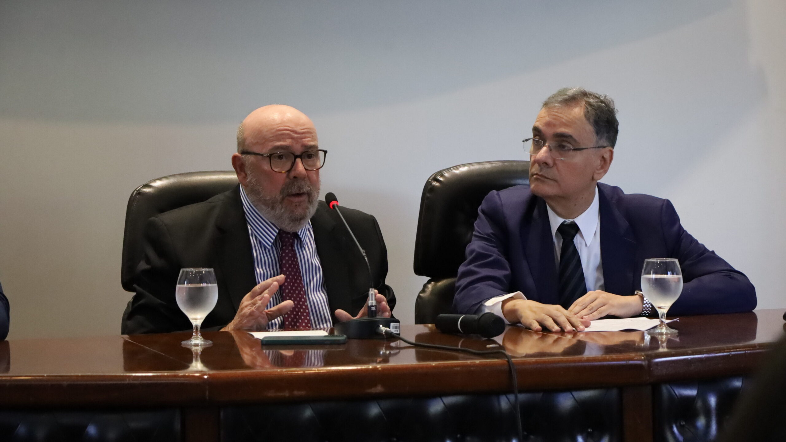 Dois homens de terno estão sentados em uma mesa de conferência com microfones e copos de água. O homem à esquerda, com barba e óculos, gesticula enquanto fala, enquanto o outro escuta atentamente.