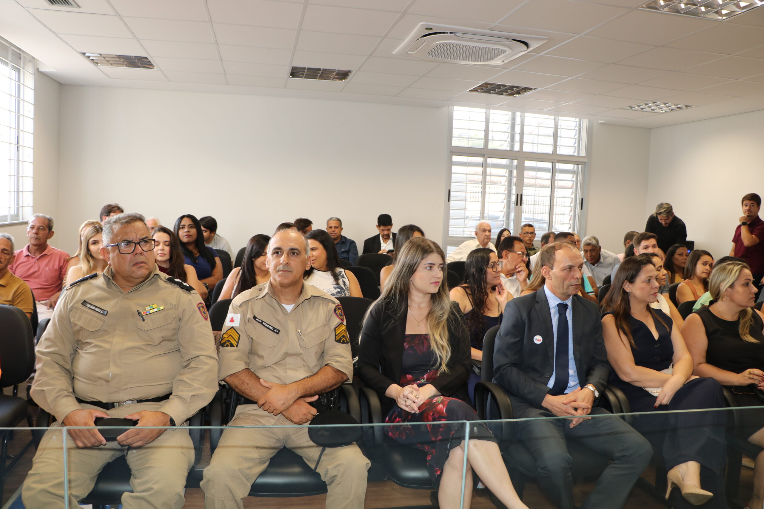 Uma plateia composta por diversas pessoas assiste a um evento em um auditório iluminado. Na primeira fileira, destacam-se dois homens em uniformes militares beges sentados ao lado de civis em trajes formais, todos voltados para a frente em atitude de escuta.