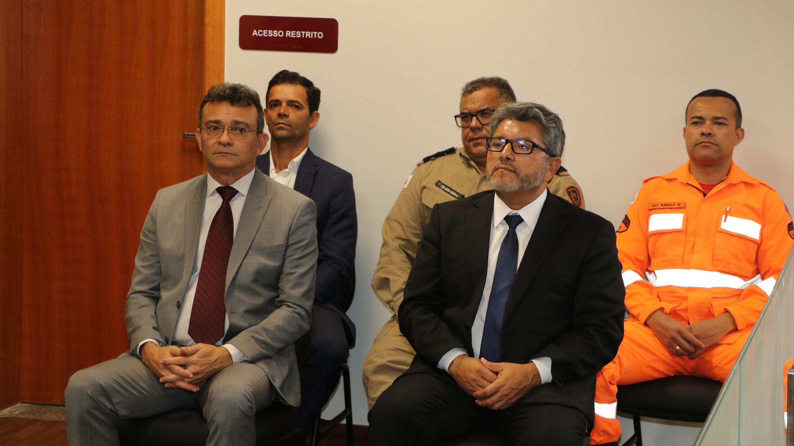 Cinco homens, dois deles vestindo uniformes de forças de segurança (um em bege e outro em laranja reflexivo), estão sentados lado a lado em uma cerimônia. Eles mantêm uma postura atenta e solene, posicionados em frente a uma porta com a placa "Acesso Restrito".