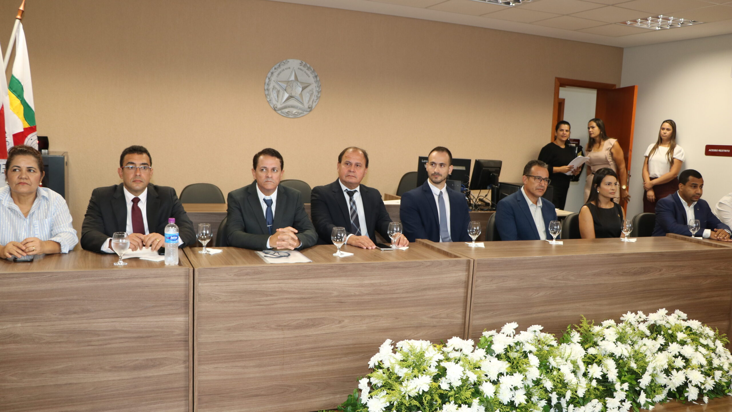Oito pessoas, entre homens e mulheres em trajes formais e casuais, estão sentadas lado a lado em uma longa mesa de madeira durante um evento institucional. Ao fundo, veem-se bandeiras e um brasão na parede, enquanto o grupo observa atentamente a cerimônia.