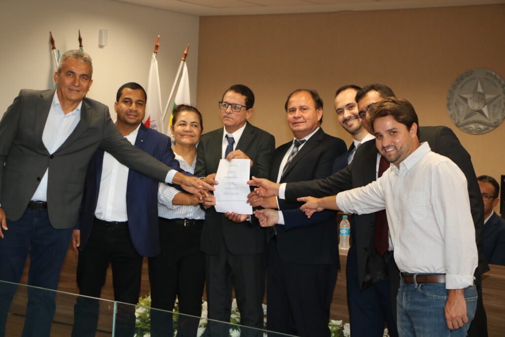 Oito pessoas, vestindo trajes formais e casuais, posam juntas segurando um documento oficial branco em um ambiente institucional. Ao fundo, veem-se bandeiras e um brasão na parede, caracterizando um ato de assinatura ou entrega solene.