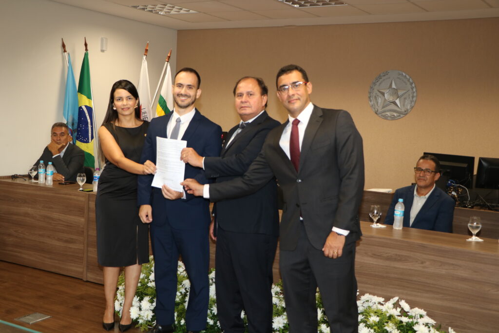 Quatro pessoas, uma mulher em vestido preto e três homens de terno, posam sorridentes em primeiro plano segurando um documento oficial. Ao fundo, outras pessoas estão sentadas em uma mesa de reunião, sob bandeiras e um brasão institucional na parede.